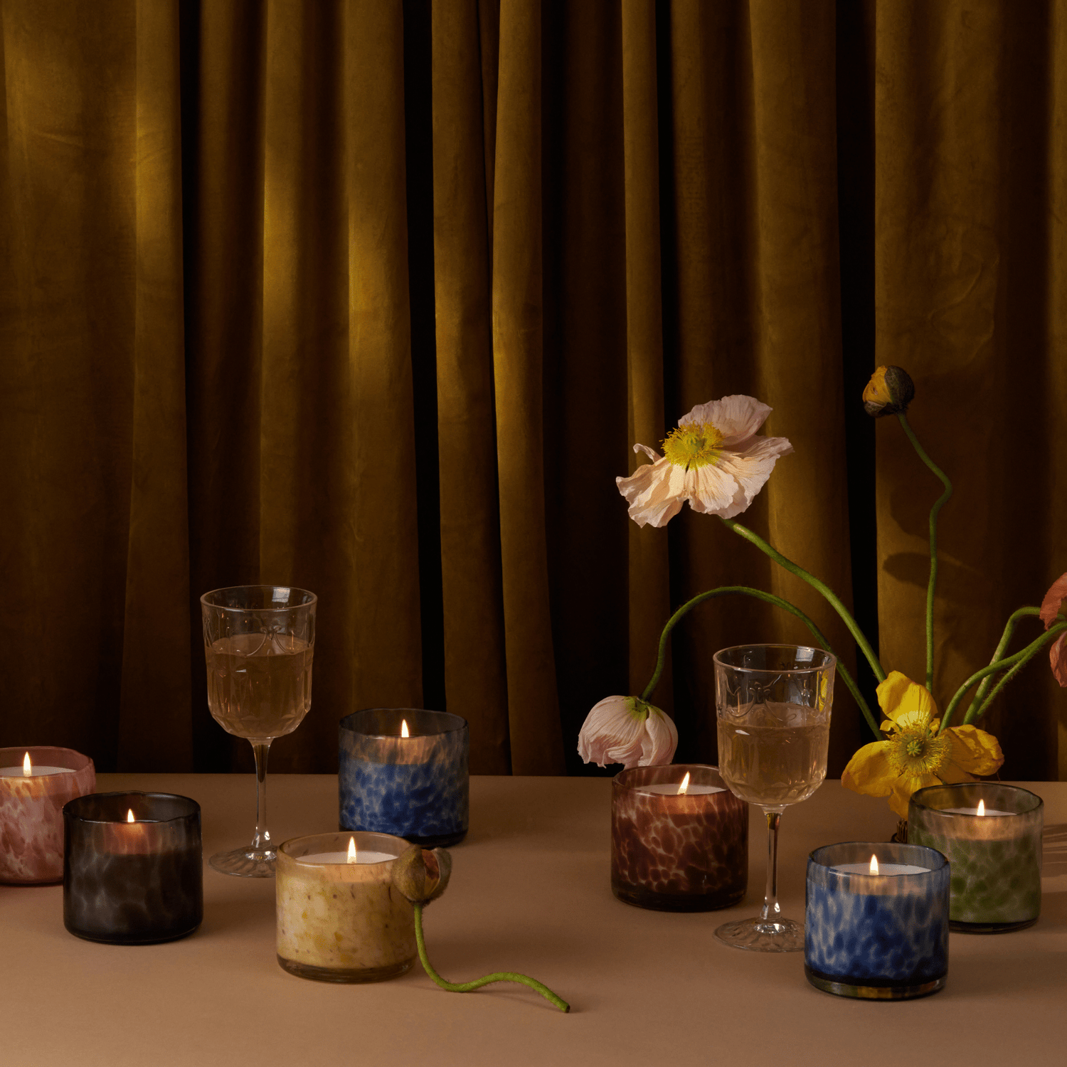 Luxe candles on a table with flowers and wine glasses and a curtain in the background