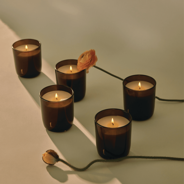 Five Firefly Signature glass candles on plain surface with two flowers and heavy shadow
