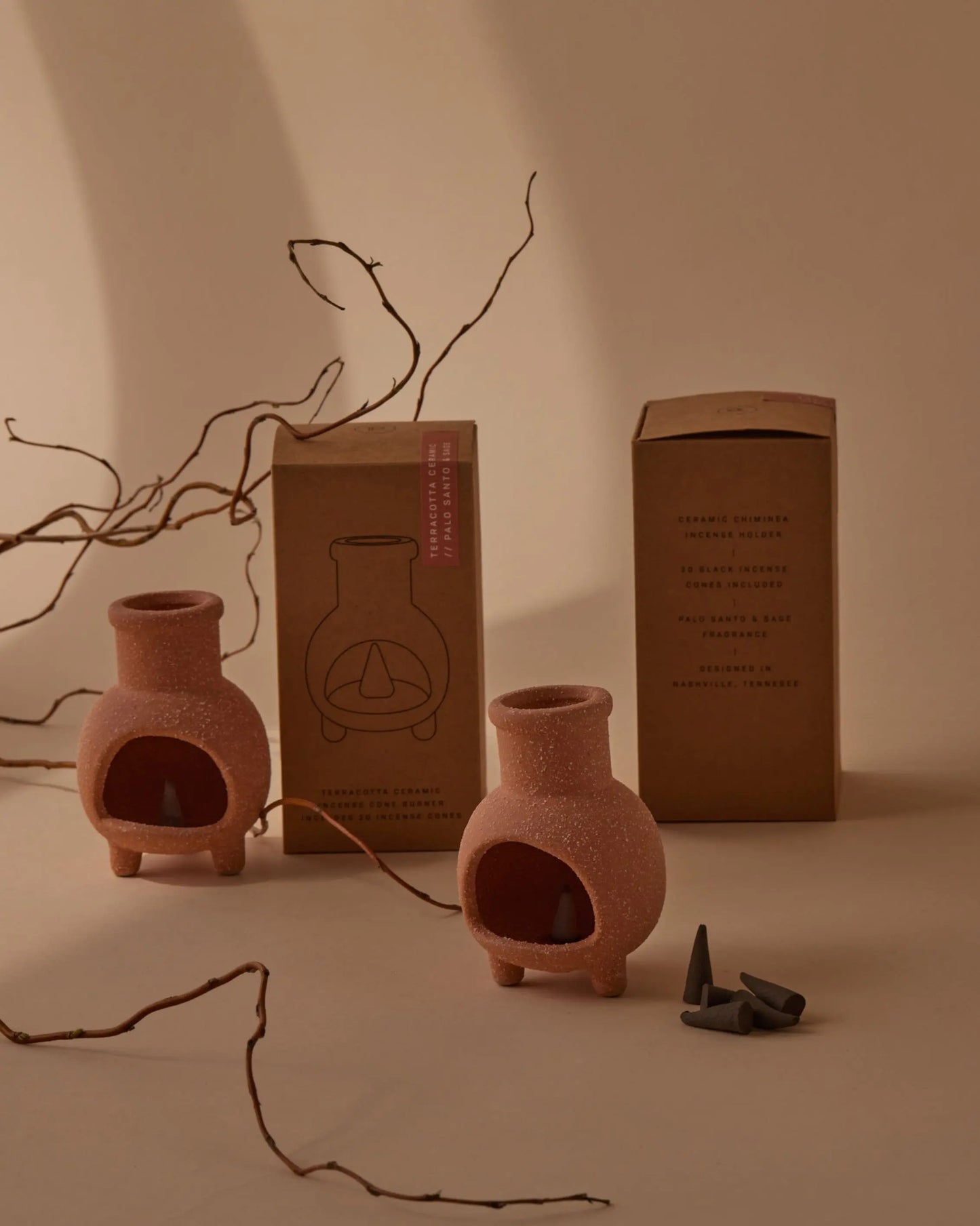 Two Ceramic Chimnea Incense Cone Holders are placed near their cardboard packaging, surrounded by incense cones and dry branches on a beige background—a stylish ceramic accessory for any space.