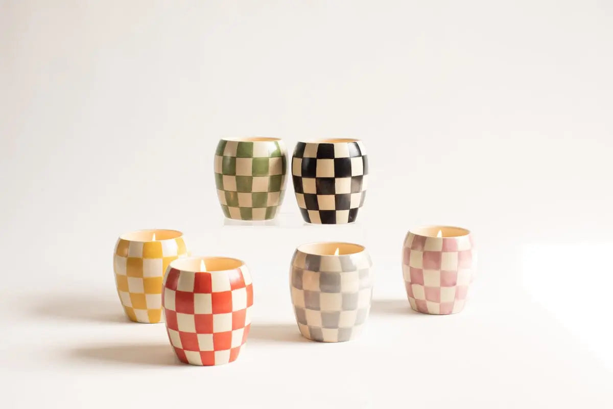 Five Checkmate 11 oz Candles - Rose + Santal, in hand-painted vessels with vibrant yellow, green, black, red, and pink checkerboard prints, are arranged on a plain white surface against a neutral background.