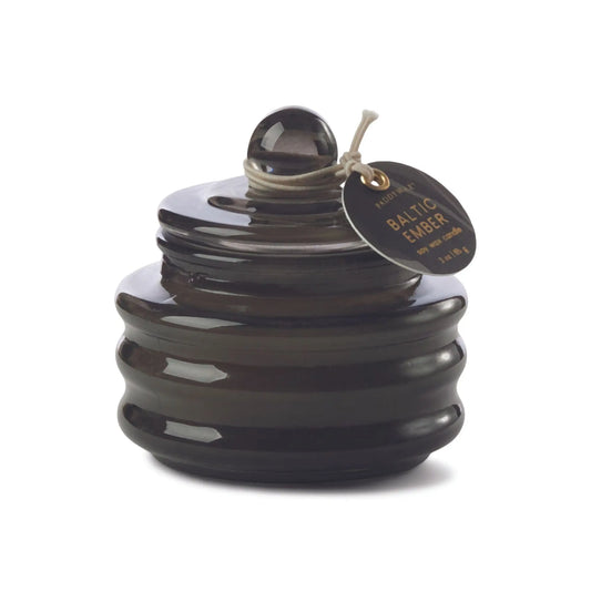 The Beam 3oz Glass Candle - Baltic Ember comes in a dark glass jar with a round lid and tag, filling your space with luxurious scents and casting a soft, ambient light.