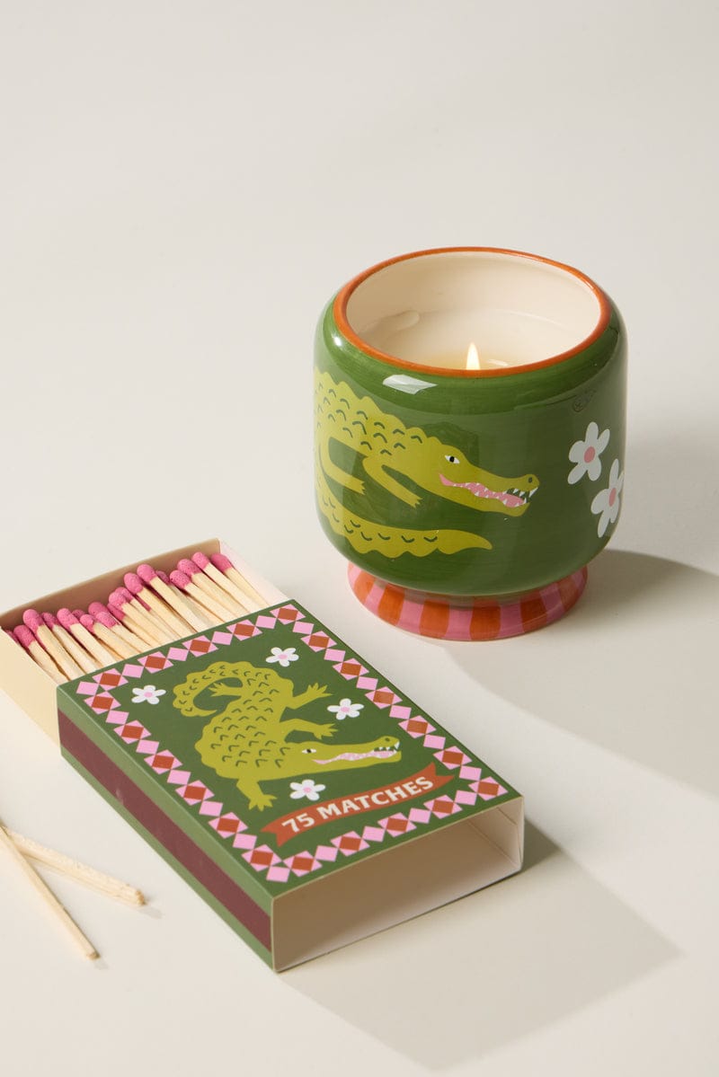 A playful green candle holder with a crocodile illustration sits next to an A Dopo "Crocodile"—75ct Boxed Matches, both featuring whimsical crocodile and floral designs, perfect for pairing with your favorite A Dopo candle.
