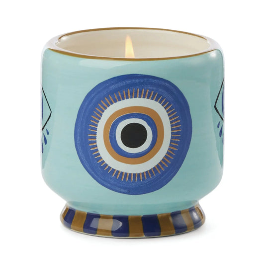 A Dopo Eye Candle - Incense & Smoke: a lit ceramic candle in a holder with hand-painted blue and gold eye accents and a striped base, shown on a white background.
