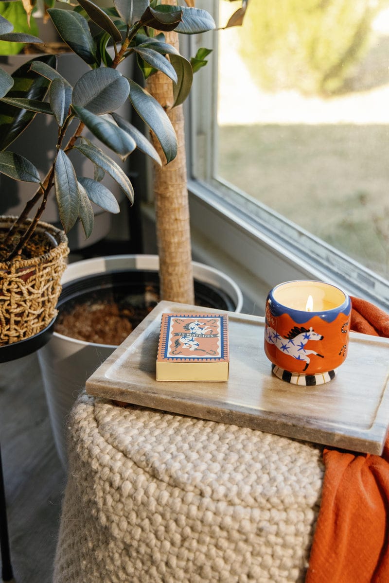 An A Dopo candle in a decorative holder and an A Dopo "Horse" - 75ct Boxed Matches rest on a marble tray beside a potted plant and woven basket near a sunlit window.