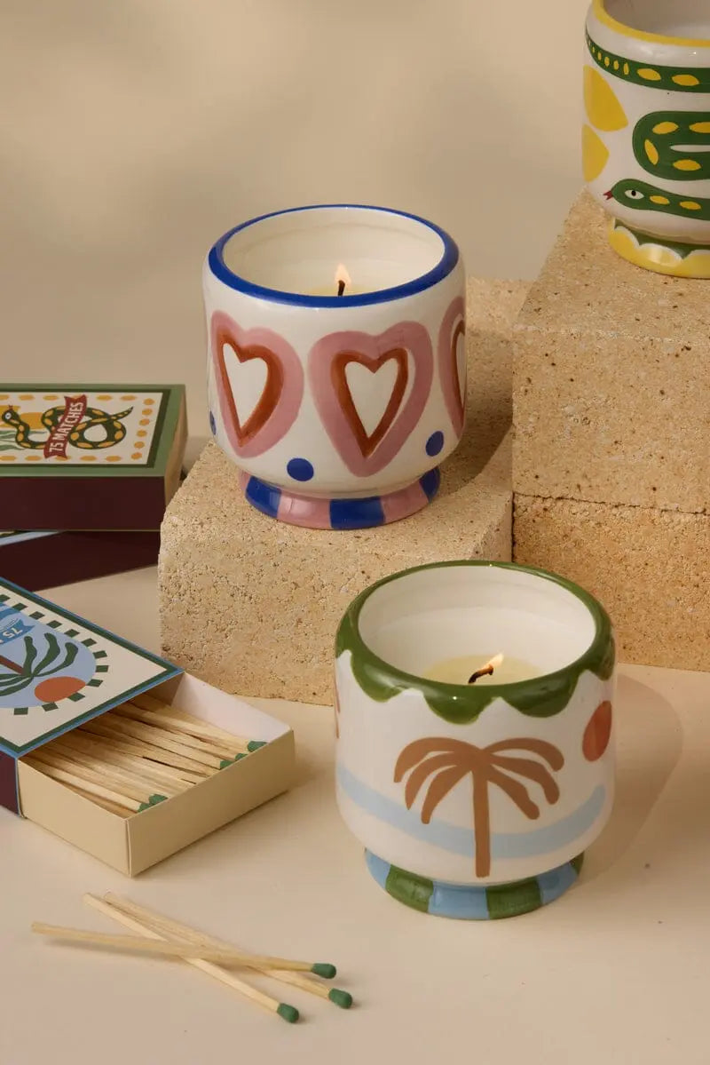 The A Dopo Hearts Candle - Rosewood Vanilla, in a painted ceramic holder with heart design, glows beside open matchboxes on beige blocks—adding bold fragrance and wanderlust-inspired style to any space.