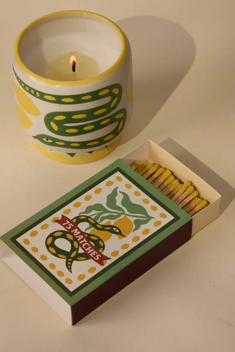 A lit A Dopo candle in a ceramic holder with a snake design, paired with the A Dopo Snake 75ct Boxed Matches—both featuring matching snake illustrations inspired by Mediterranean travels.