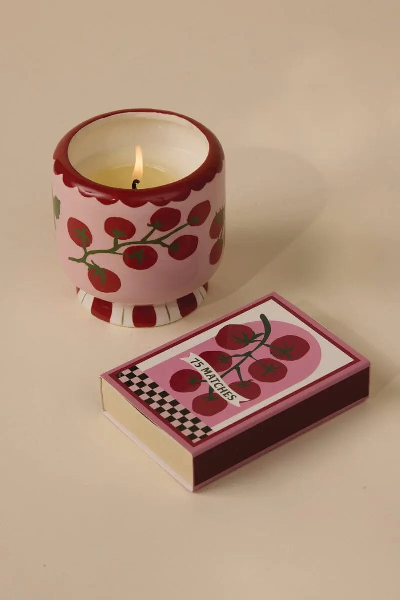 A pink ceramic candle holder with red tomato designs sits next to an A Dopo Tomato Vine 75ct boxed matches, creating a charming Mediterranean vibe on the beige surface.