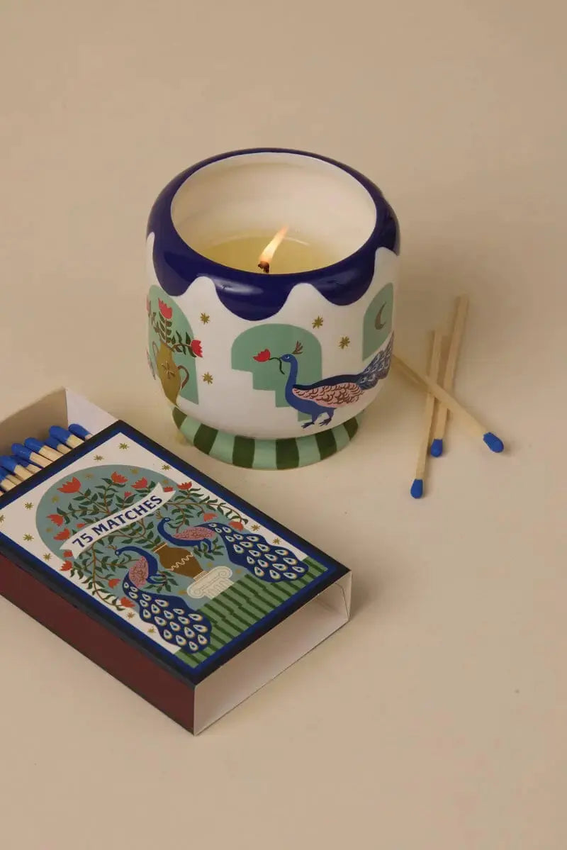 A Dopo Peacock 75ct Boxed Matches, with three matches displayed outside the open box, rest beside a lit candle in a decorative animal-themed holder on a beige background.