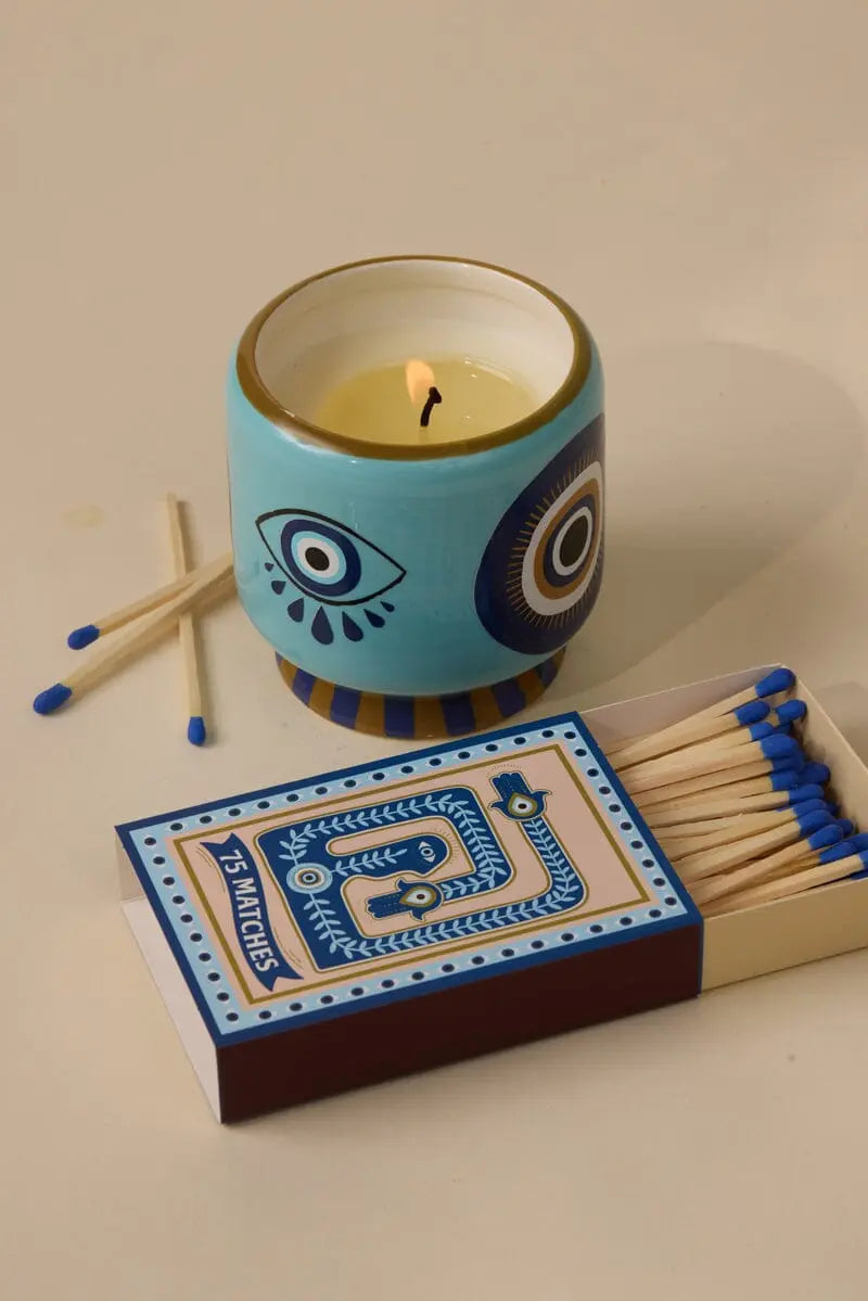 A lit candle in a blue ceramic holder with eye designs sits next to the A Dopo Eye 75ct boxed matches, some scattered nearby—a stylish touch for your candle accessories collection.