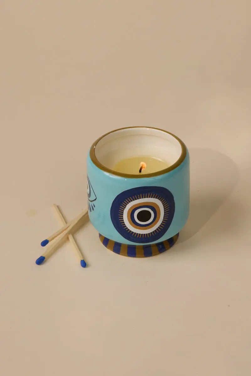 The A Dopo Eye Candle - Incense & Smoke features a lit ceramic holder with hand-painted eye details in blue, set beside three blue-tipped matches on a beige surface.