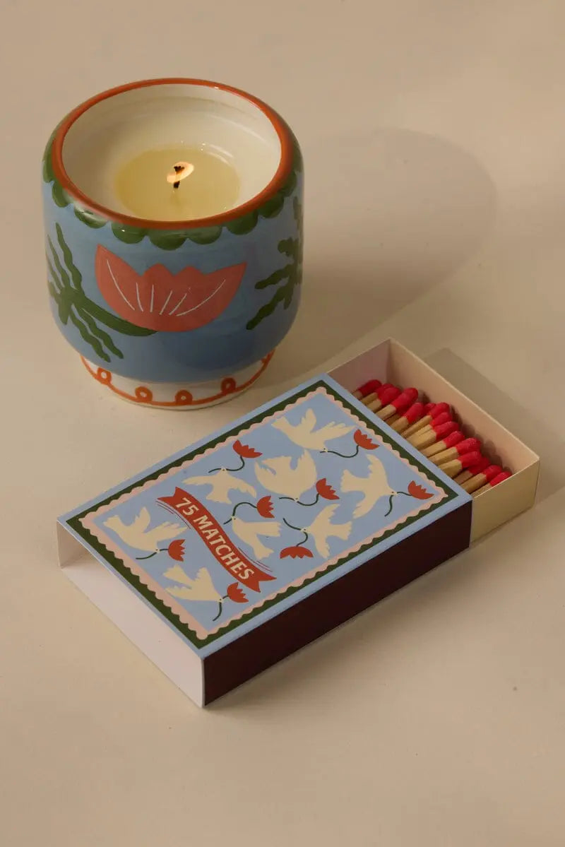 An A Dopo candle in a decorative holder sits beside an open box of A Dopo Flower - 75ct Boxed Matches, their red tips standing out against the beige surface.