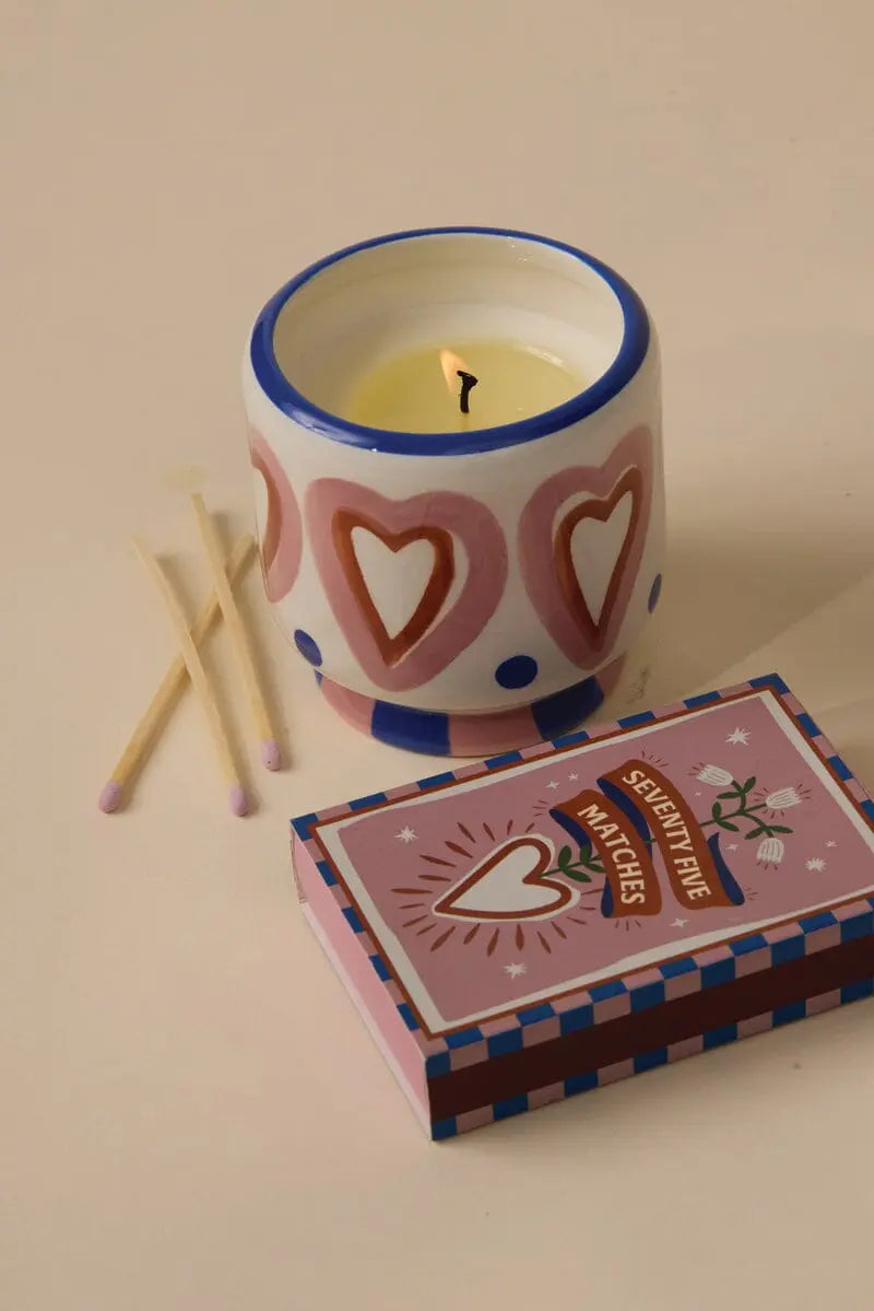 A glowing candle in a heart-patterned holder sits beside the A Dopo Hearts - 75ct Boxed Matches and three safety matches on a beige surface, evoking Mediterranean travel keepsakes.