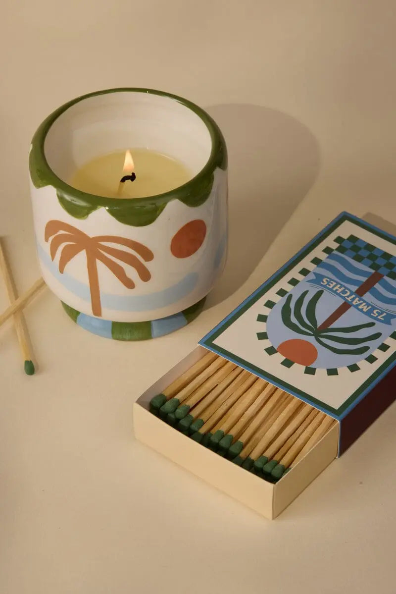 A Dopo Palm Tree 75ct boxed matches sit next to a decorative candle in a ceramic holder with a palm tree motif on a beige surface, evoking memories of Mediterranean travels.