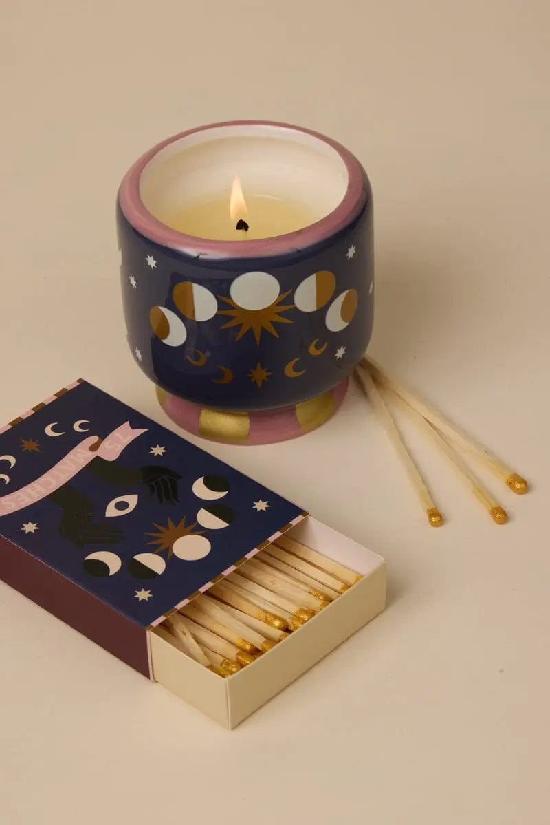The A Dopo Moon Phases Candle - Jasmine & Rose sits in a blue, pink, and gold holder beside an open moon-and-star matchbox—its bold scent evokes Mediterranean travels, with matches scattered nearby.