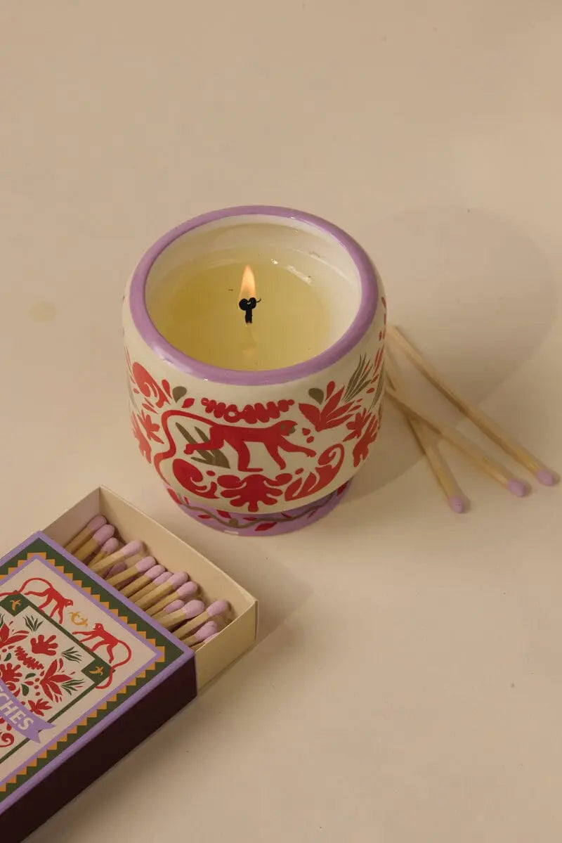 A lit A Dopo candle in a decorative holder sits beside an A Dopo Jungle – 75ct Boxed Matches and three loose matches on a beige surface, evoking memories of Mediterranean travels.