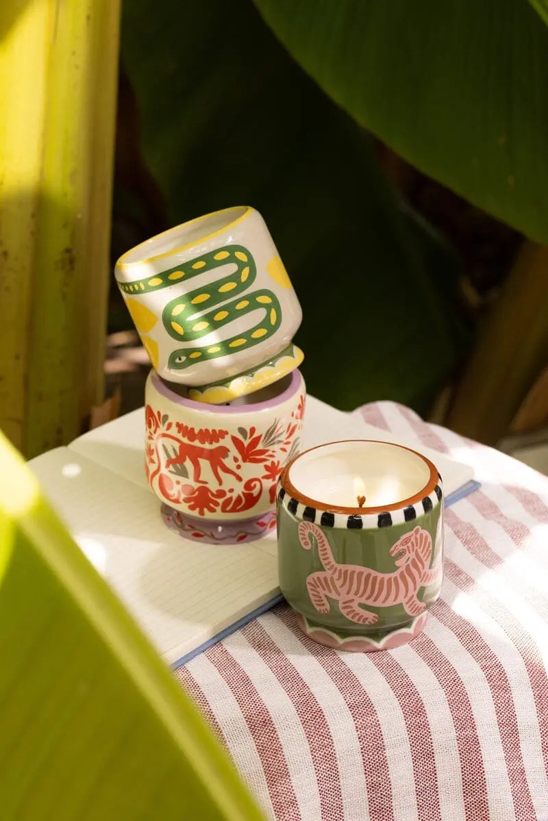 The A Dopo Jungle Candle - Coconut & Amber sits lit in a hand-painted ceramic holder on a striped cloth beside patterned holders and a notebook, large green leaves framing this serene scene.