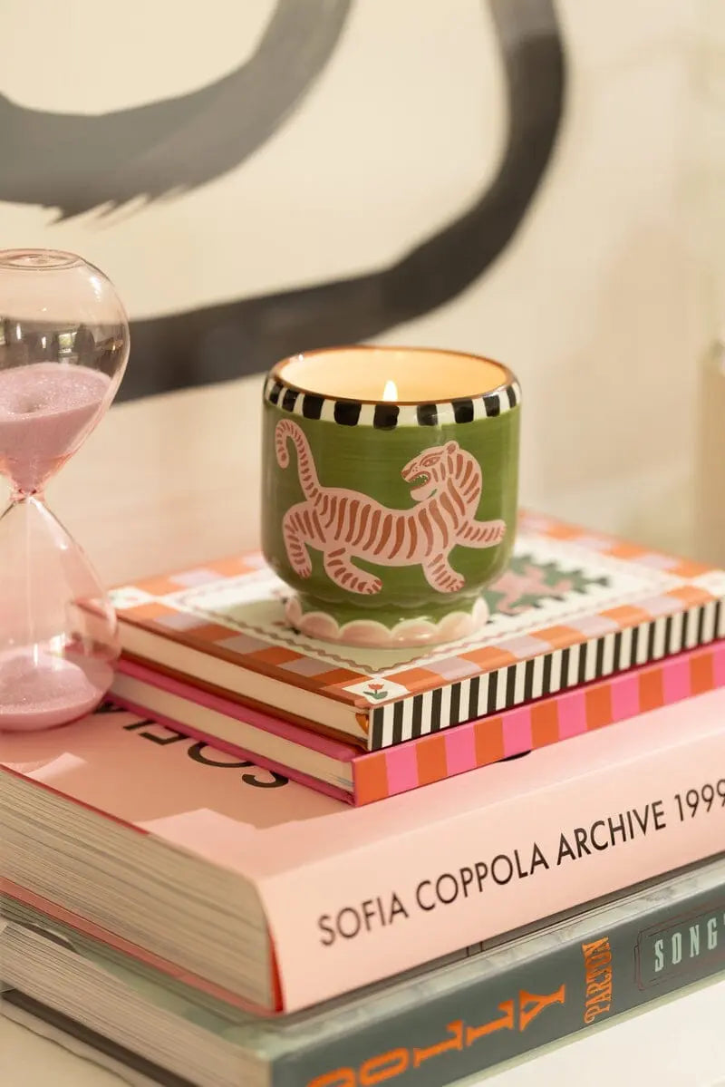 A Dopo Tiger Candle - Black Cedar & Fig, in a green ceramic holder with a white tiger design, sits atop stacked books including SOFIA COPPOLA ARCHIVE 1999–2023, its bold scent filling the air beside a pink hourglass.