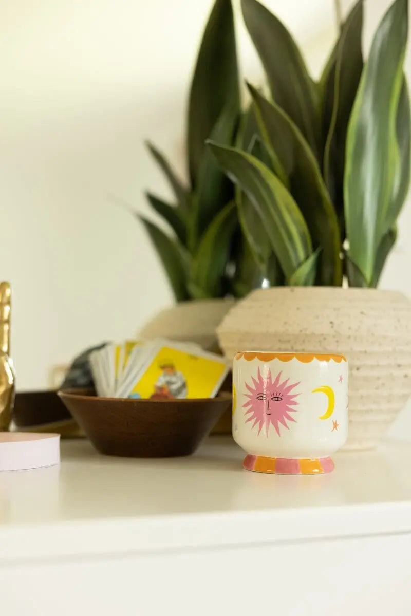 A white table with a potted plant, a ceramic mug, a wooden bowl holding cards, a gold ornament, and the A Dopo Sun Candle - Orange Blossom.