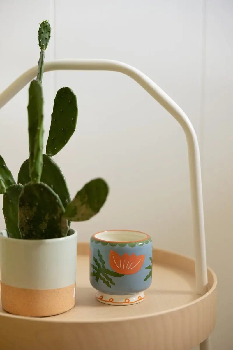 The A Dopo Flower Candle - Cactus Flower sits next to a blue cup with an orange floral pattern and hand-painted details on a light wooden tray with a white metal handle.