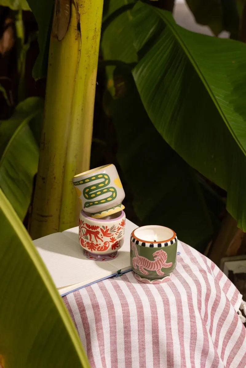 The A Dopo Snake Candle in Wild Lemongrass sits atop a striped cloth next to a notebook, framed by lush green banana leaves.