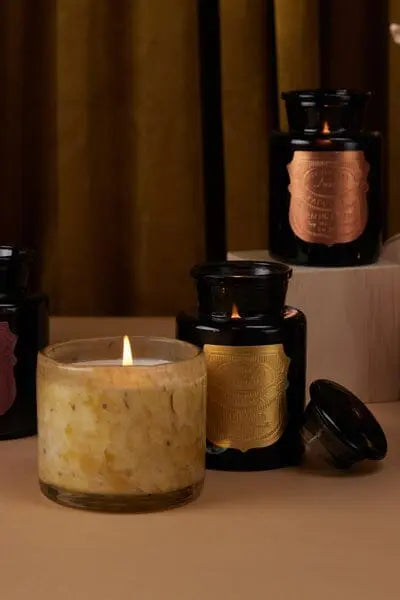 Three inky black Apothecary Noir - Palo Santo 8oz candles with metallic labels are displayed; one light-colored Palo Santo candle is lit in a clear, textured holder in the foreground.