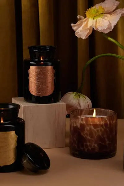 A lit Apothecary Noir - Baltic Ember 8 oz candle in marbled glass sits beside two black glass Apothecary Noir jars with gold labels on a beige surface, with pink flowers in the background.