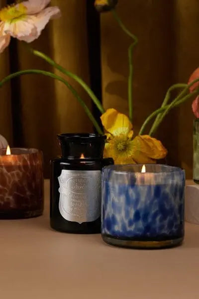 Three decorative candles, including the Apothecary Noir - Black Fig 8oz in a dark glass jar with a silver label and two patterned glass holders, are arranged on a surface with yellow flowers in the background.