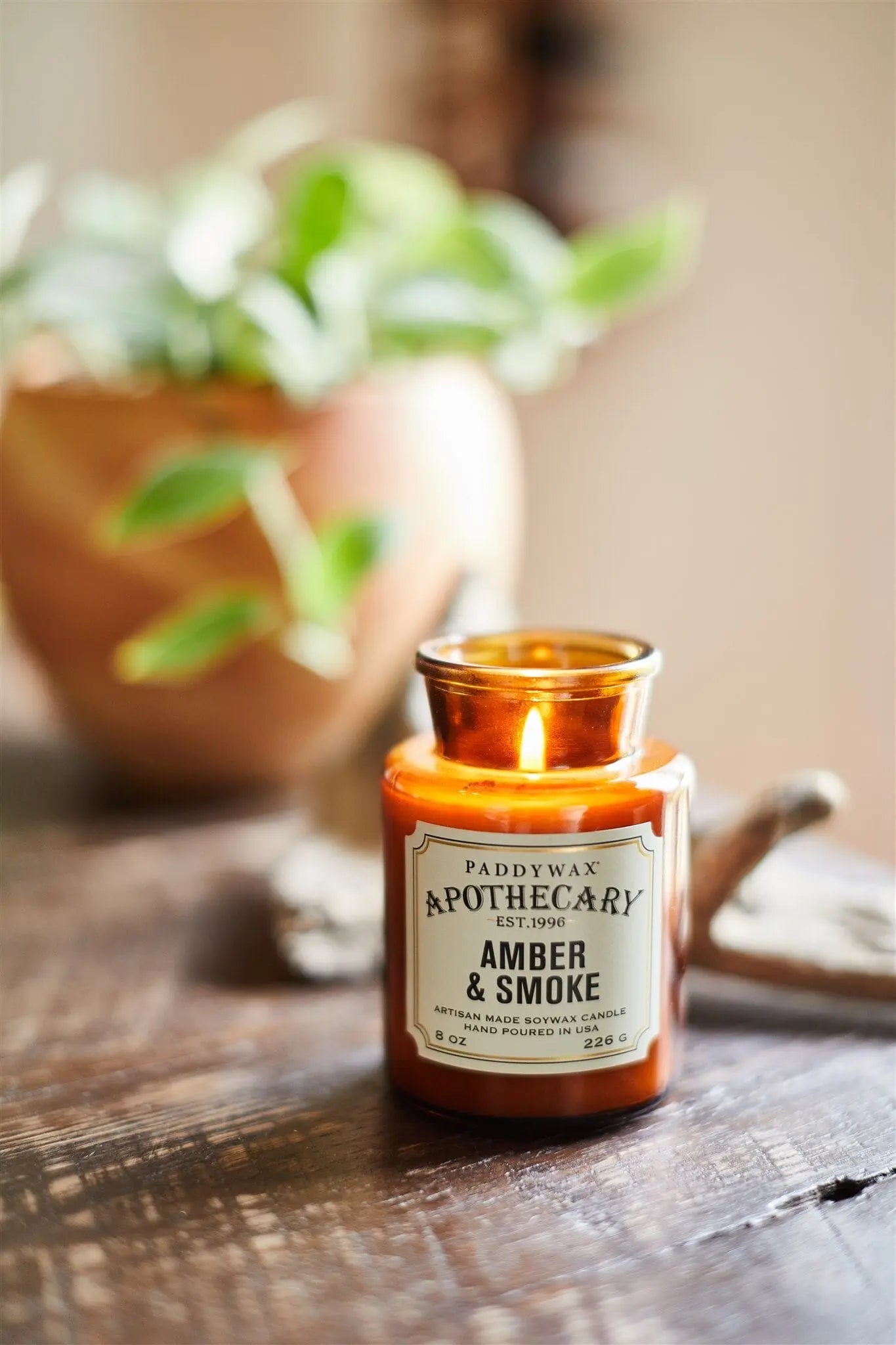 An Apothecary 8 oz Candle - Amber + Smoke is lit in its amber glass jar on a wooden surface, with a potted plant softly blurred in the background.
