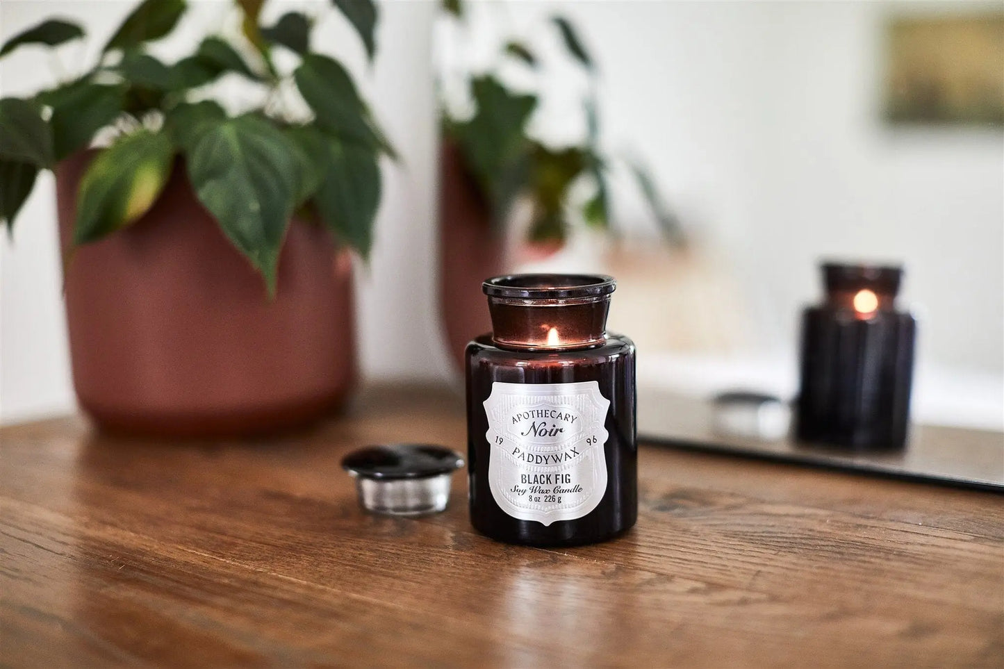 An Apothecary Noir - Black Fig 8oz luxury candle in a black glass vessel rests on a wooden surface near a potted plant and mirror, with its lid beside it, adding an elegant touch to the decor.