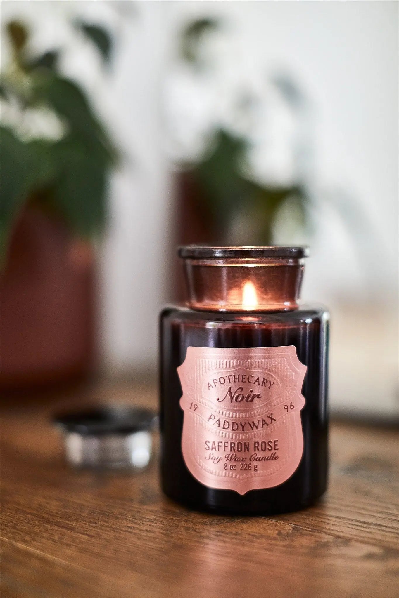 An Apothecary Noir - Saffron Rose 8oz luxury candle burns on a wooden surface with its lid off, while blurred plants provide a soft background.