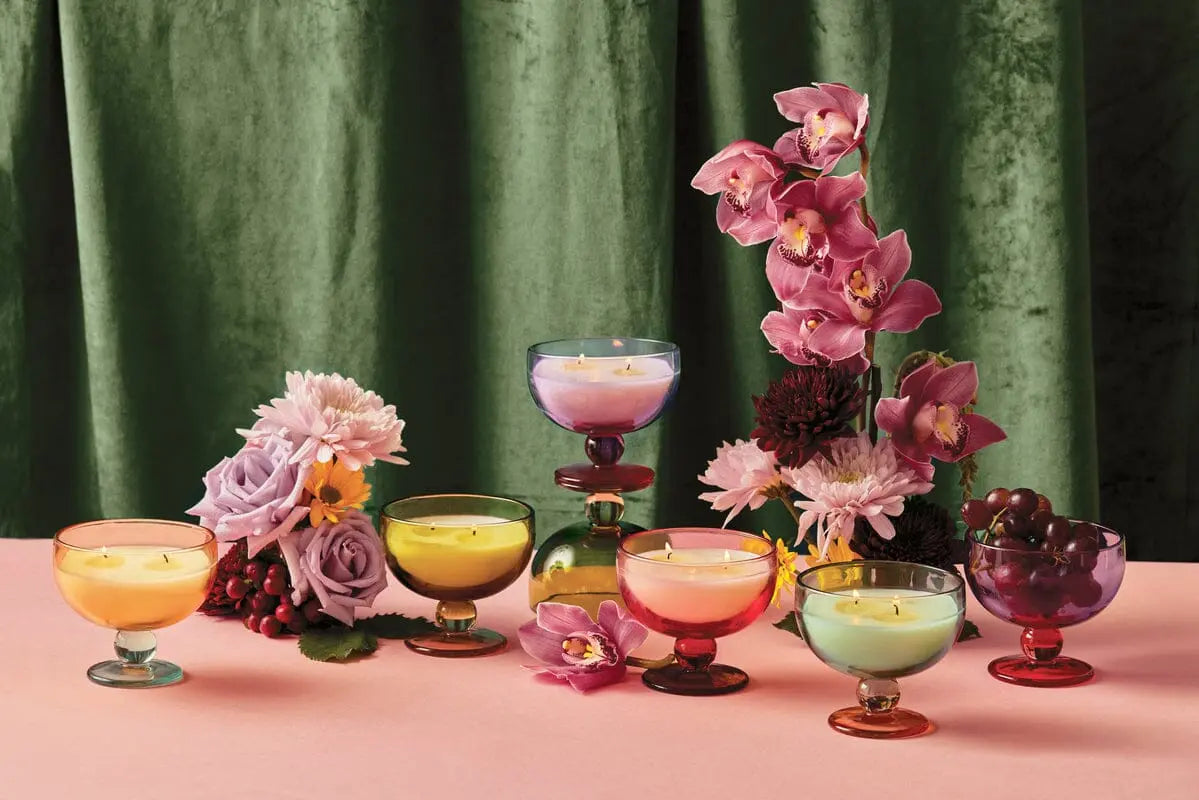 Six colorful vintage glassware candle holders with Aura 6 oz. Candle – Misted Lime are lit and arranged on a pink surface with assorted flowers, grapes, and a vibrant misted lime accent, set against a green velvet curtain.
