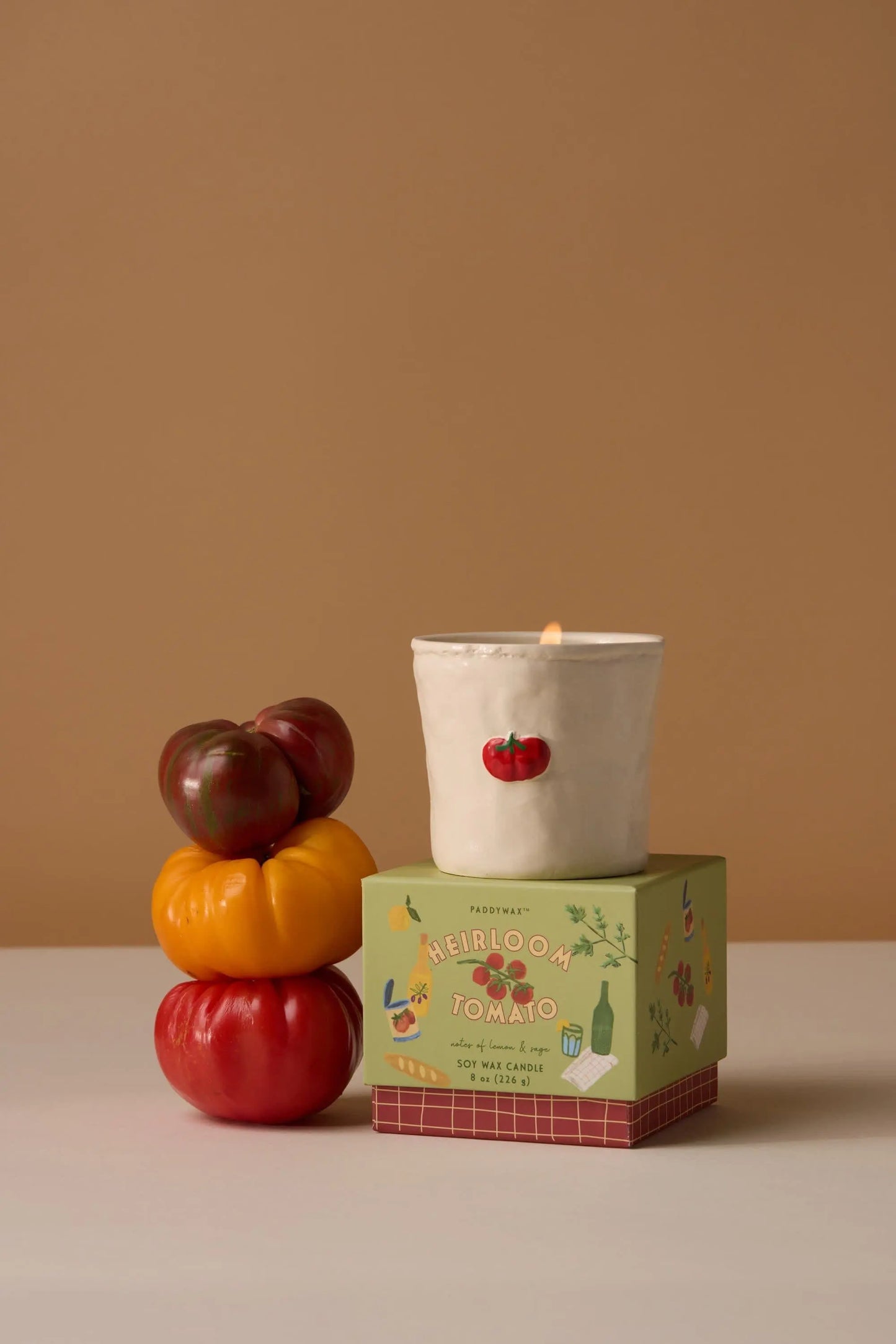 A lit Bistro Candle - Heirloom Tomato from Paddywax, featuring hand-painted food icons, sits on a green box beside a stack of three heirloom tomatoes in red, yellow, and purple.