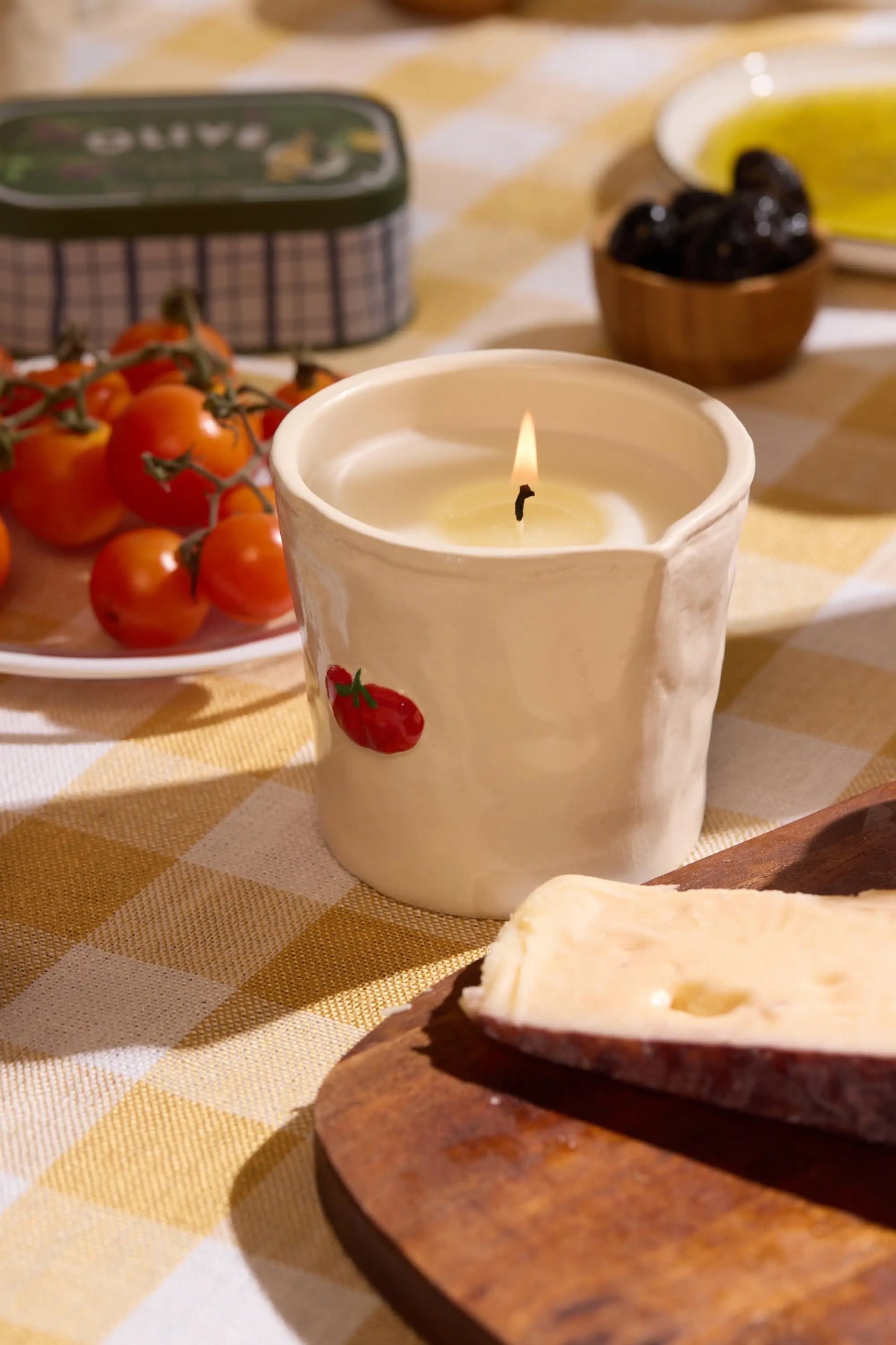 The Bistro Candle - Heirloom Tomato, set in a ceramic holder with hand-painted food icons and an heirloom tomato motif, rests on a checkered tablecloth beside cherry tomatoes, cheese, olives, and a tin.