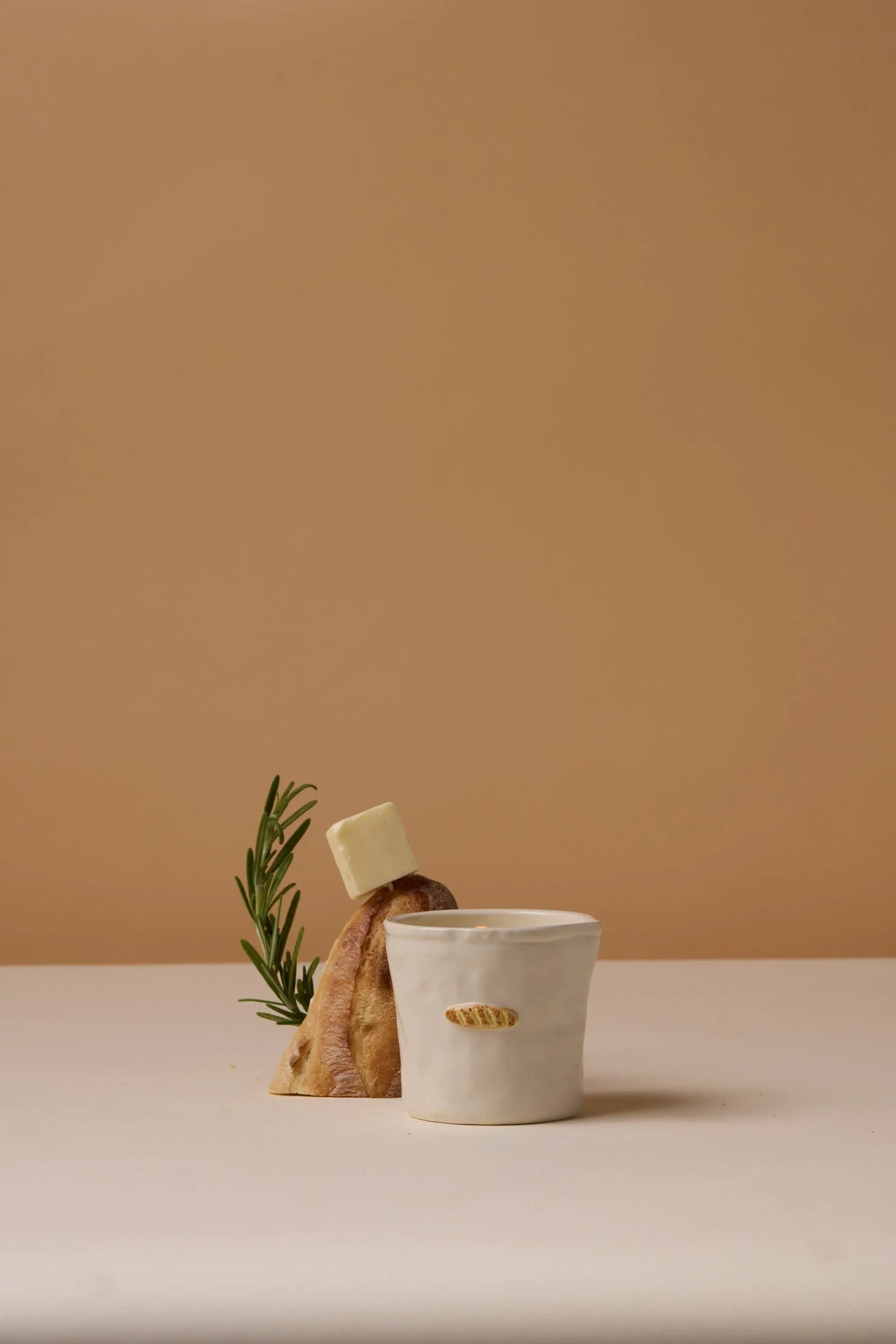 A Bistro Candle - Baguette, designed to look like a slice of bread with rosemary and butter, is set behind a white ceramic cup on a light surface against a beige background.