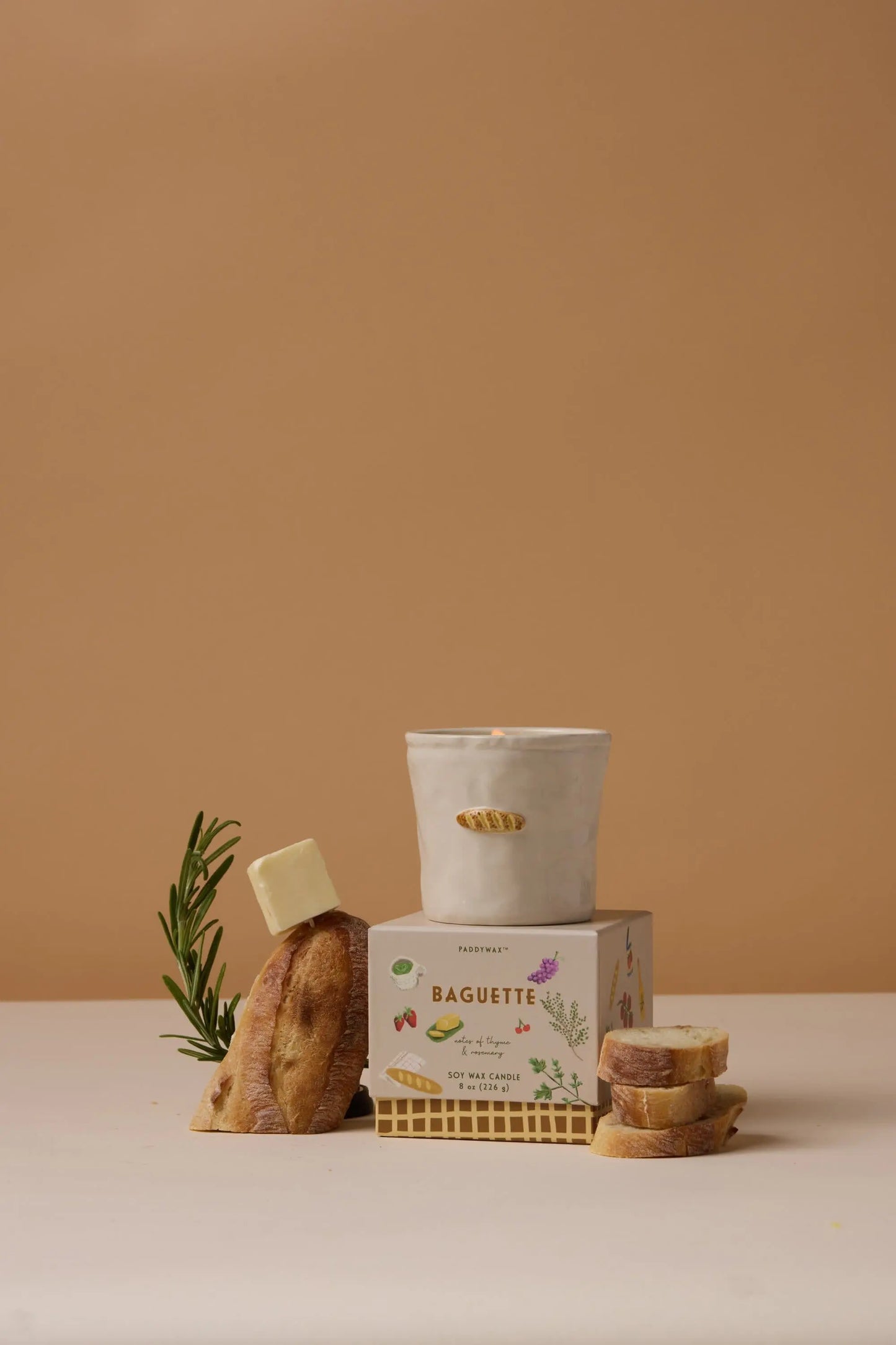 The Bistro Candle - Baguette sits atop a box labeled Baguette, surrounded by sliced baguette, rosemary, and a cube of butter—showcasing culinary culture on a beige background.