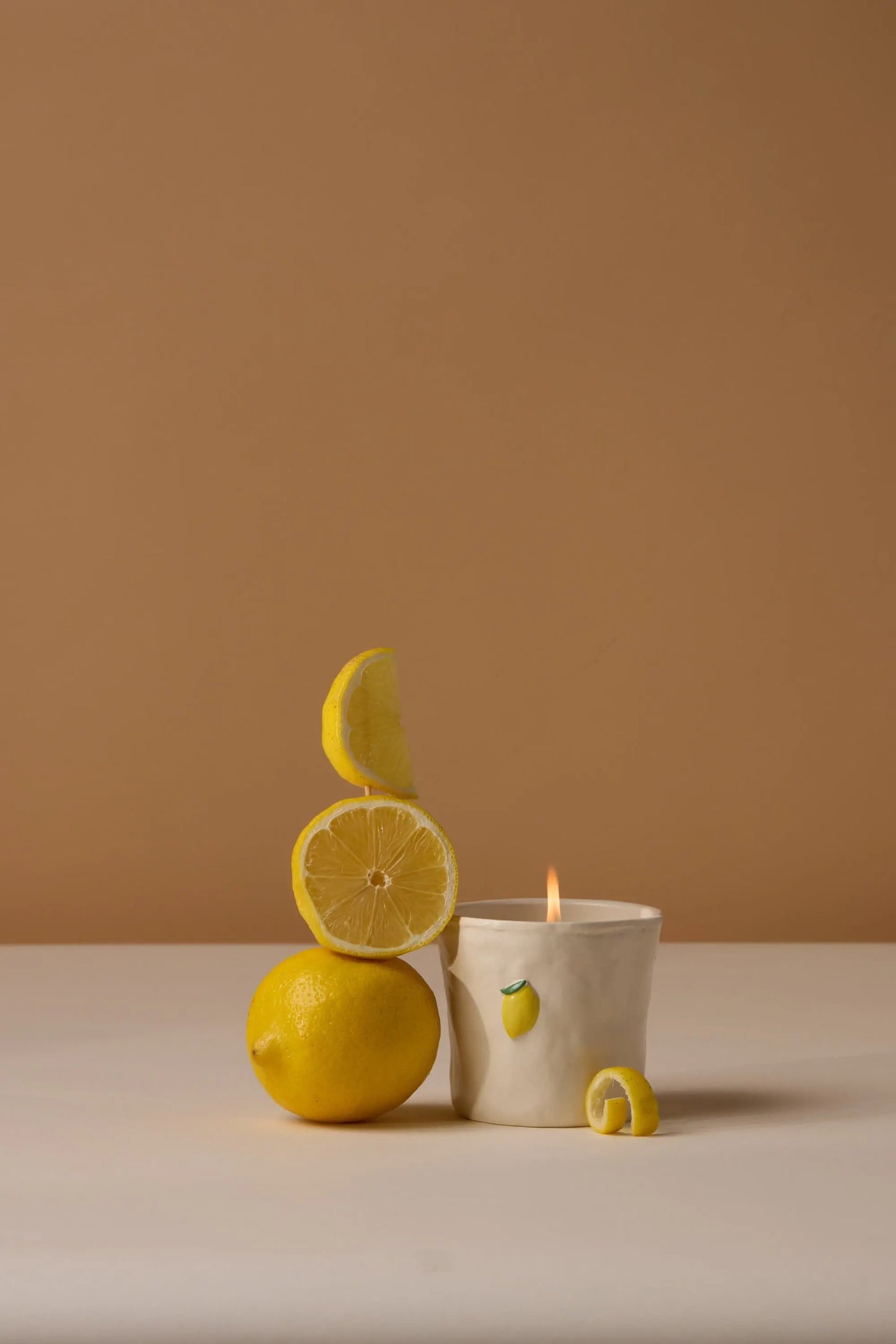 The Bistro Candle - Meyer Lemon, lit in its ceramic holder with a Meyer Lemon motif, sits beside a whole lemon, slices, and peel on a beige surface against a brown background.