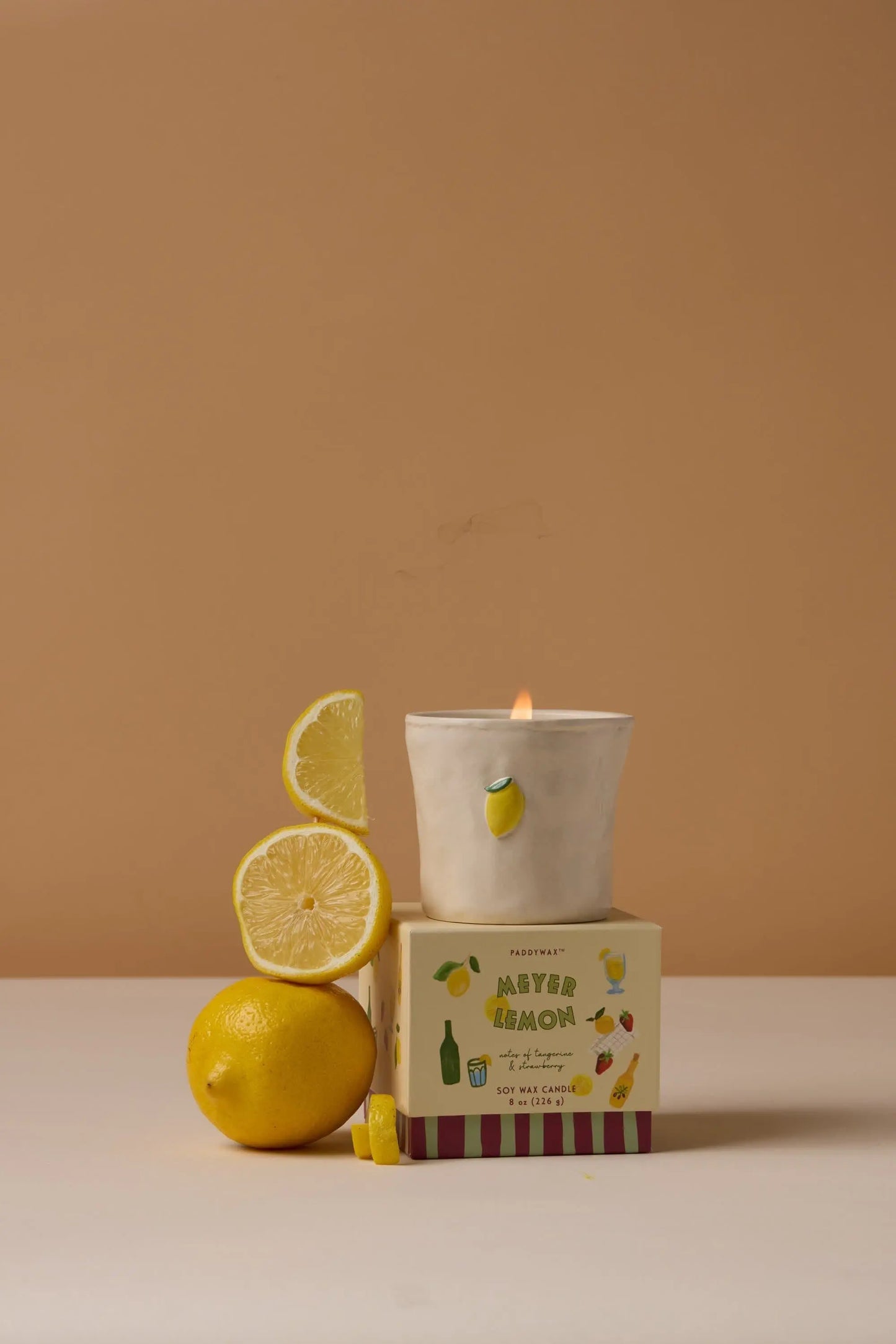 The Bistro Candle - Meyer Lemon from the Bistro Collection sits on an After Lemon box next to a whole lemon and two lemon slices, against a beige background.