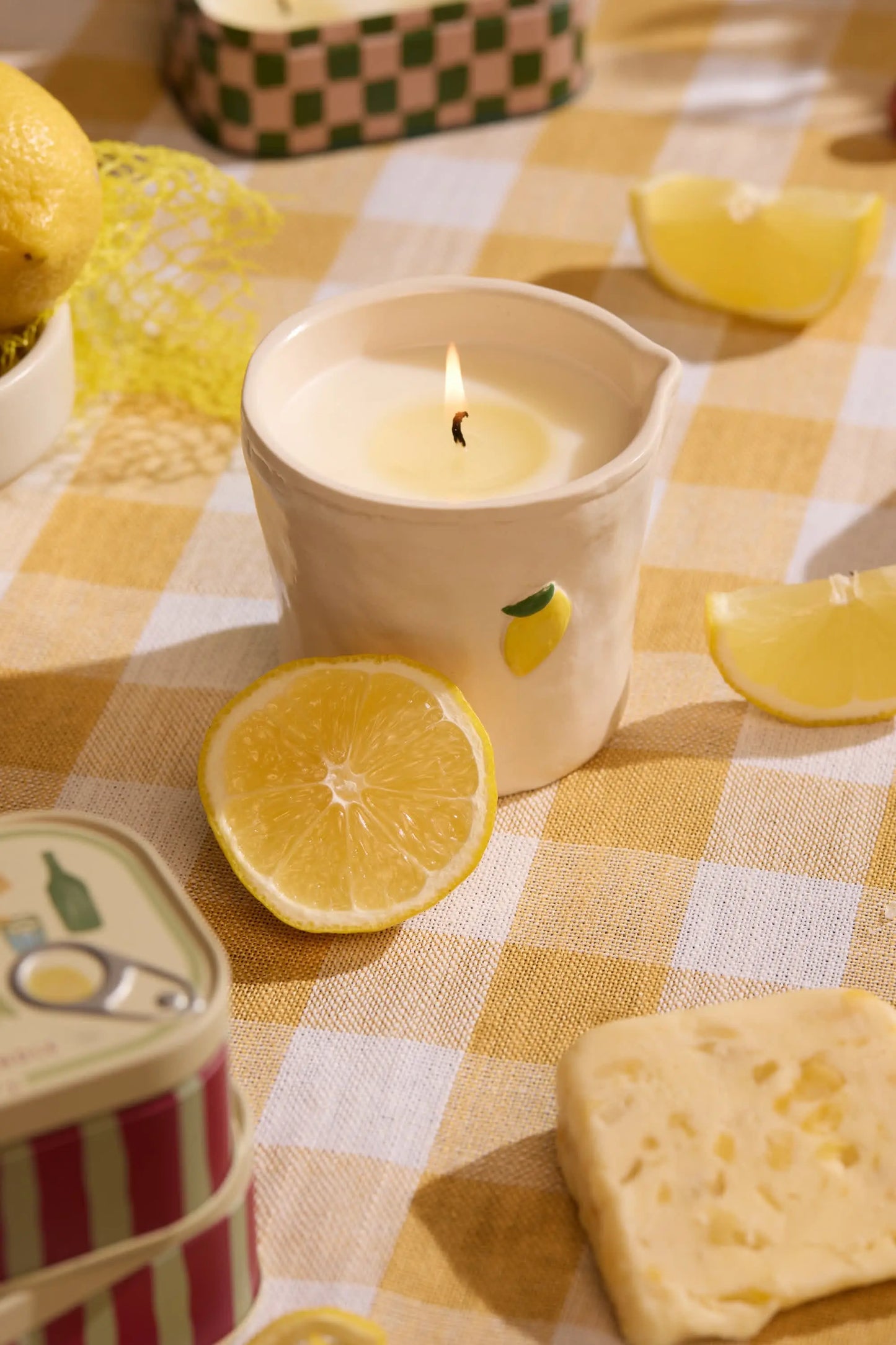 A lit Bistro Candle - Meyer Lemon from the Bistro Collection sits on a yellow checkered tablecloth, surrounded by lemon slices, cheese, and tins.
