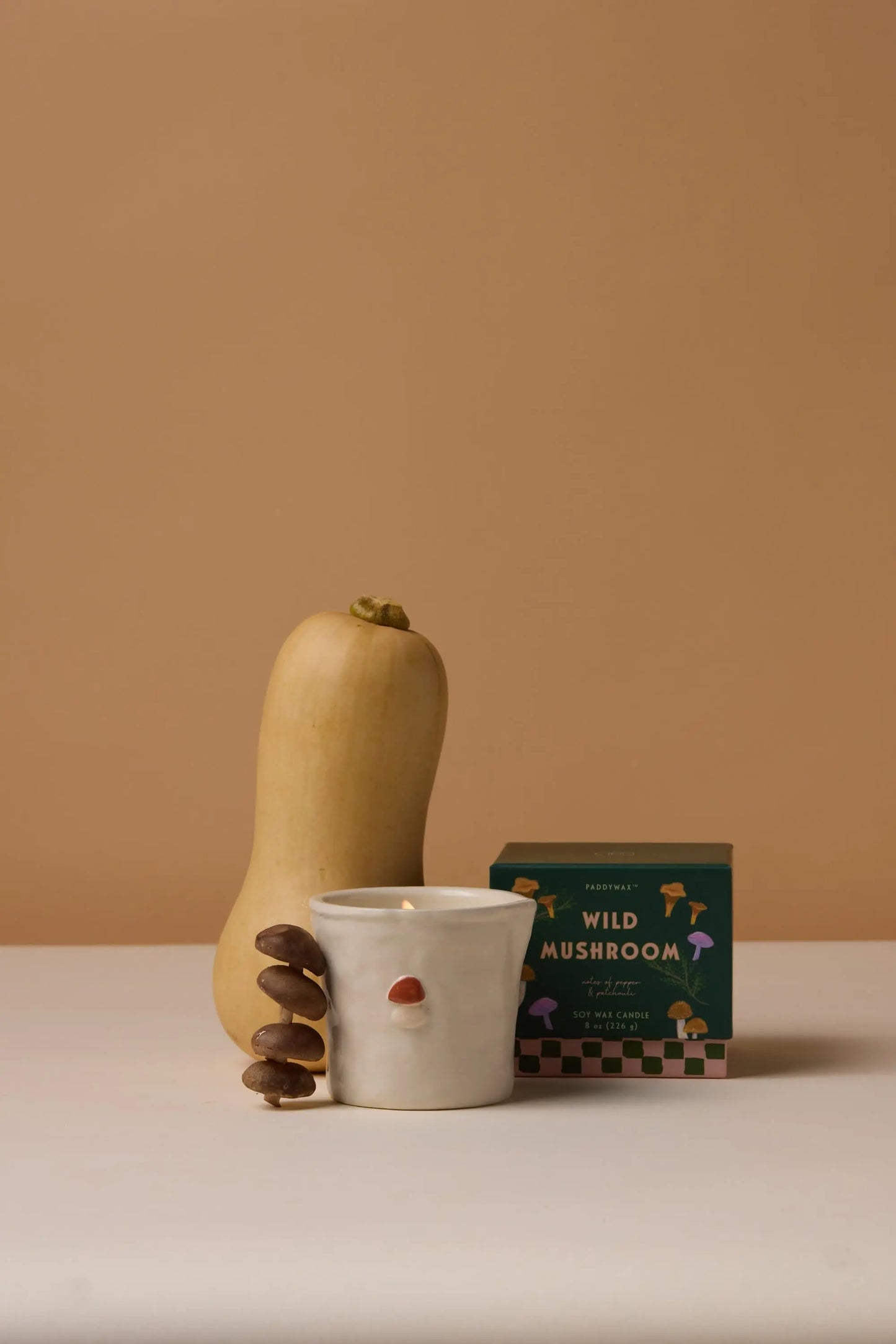 A butternut squash, a cluster of mushrooms, and the Bistro Candle - Wild Mushroom in its green box are arranged on a white surface against a beige background.