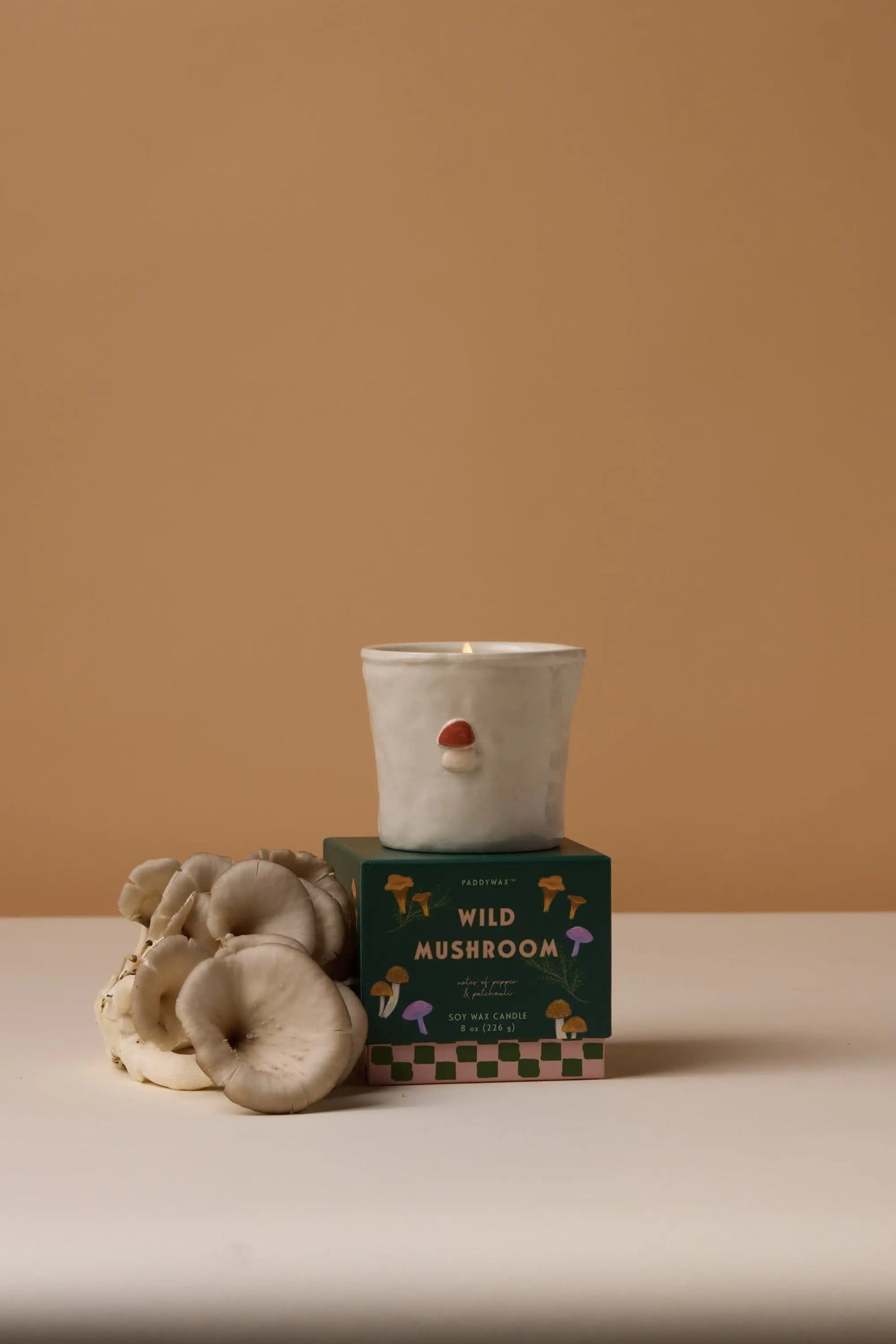 The Bistro Candle - Wild Mushroom, a white ceramic candle holder with a nose design, sits atop its green box beside white mushrooms, bringing aromatic fragrance to the beige backdrop.