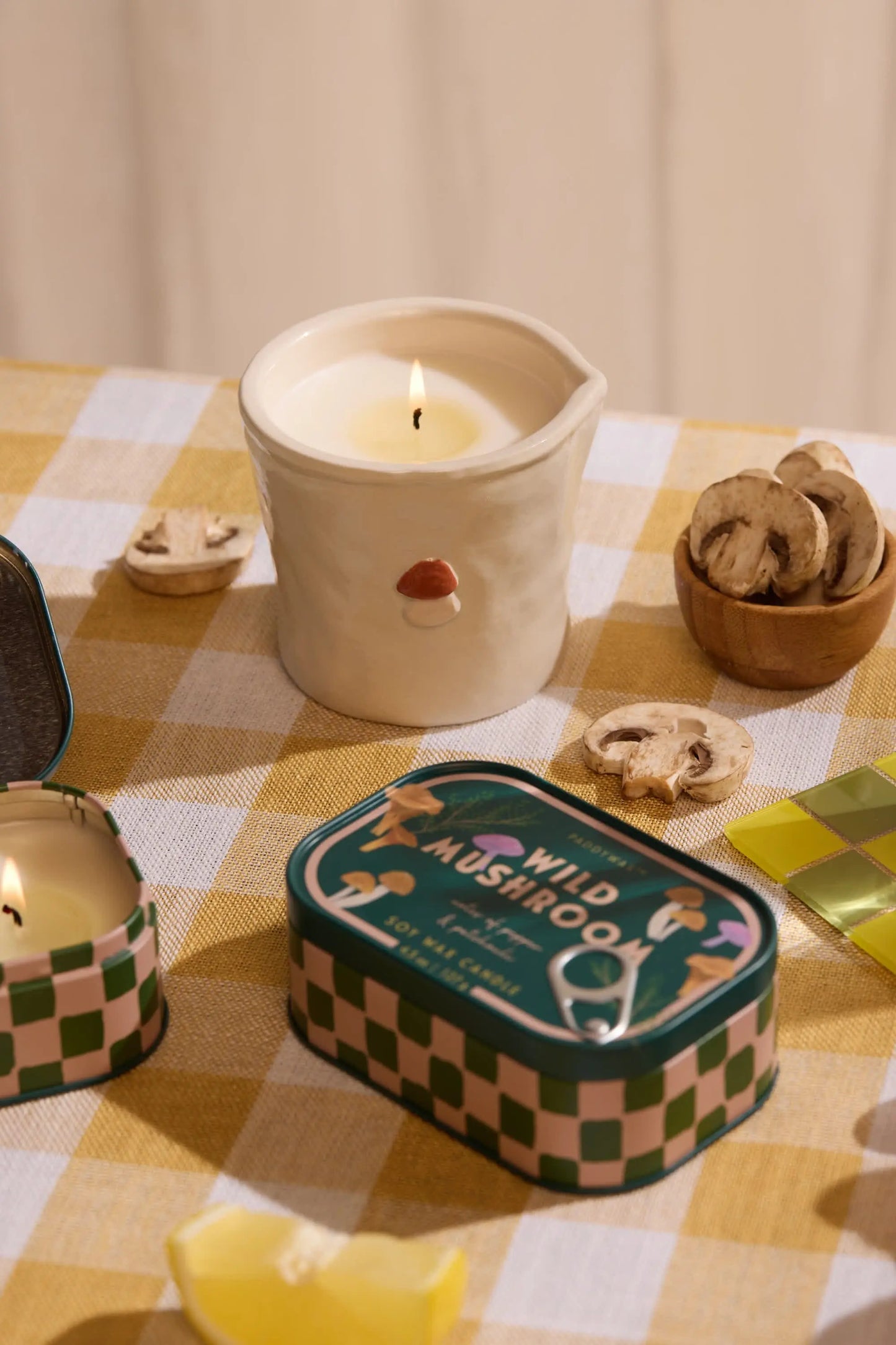 The Bistro Candle - Wild Mushroom from the Bistro Collection, with a mushroom design, sits on a yellow checkered tablecloth among dried mushrooms and a Wild Mushroom tin, filling the room with its aromatic fragrance.