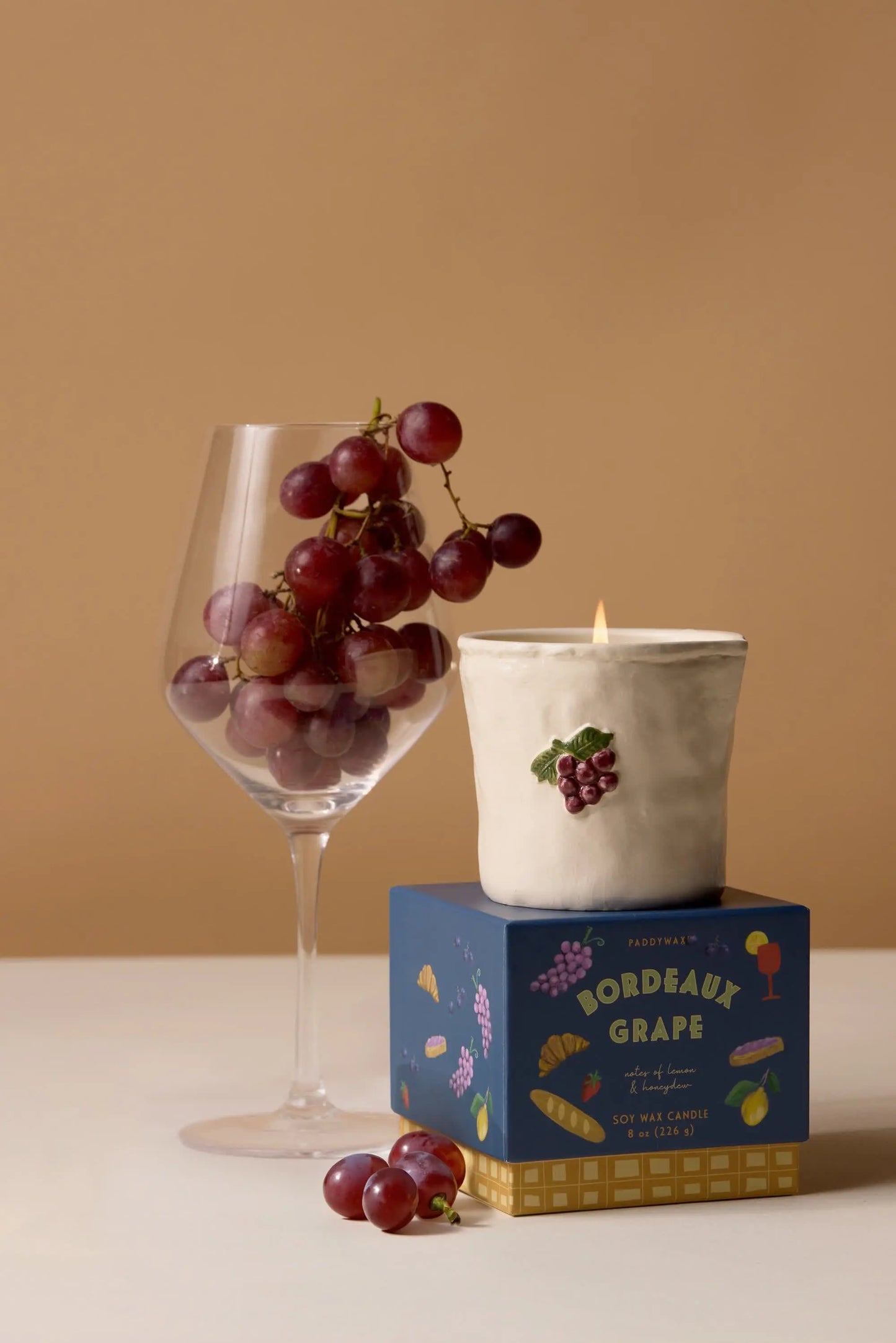 A wine glass with red grapes sits next to the Bistro Candle - Bordeaux Grape in a ceramic holder, both arranged on a blue box, capturing the essence of culinary culture.