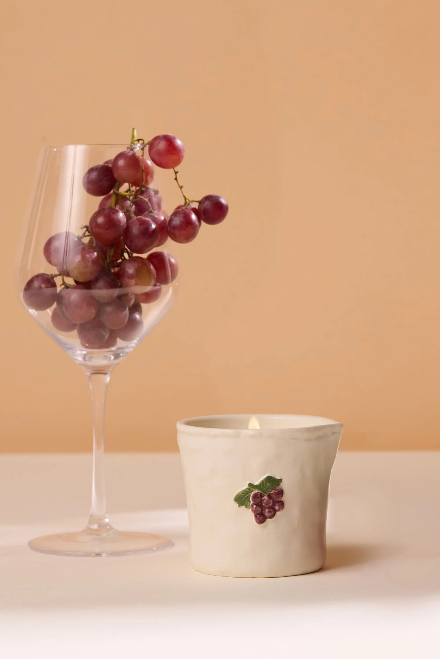 A bunch of red grapes sits in a wine glass beside the Bistro Candle - Bordeaux Grape, which features a grape motif cup, all set against a beige background to evoke a sense of culinary culture.