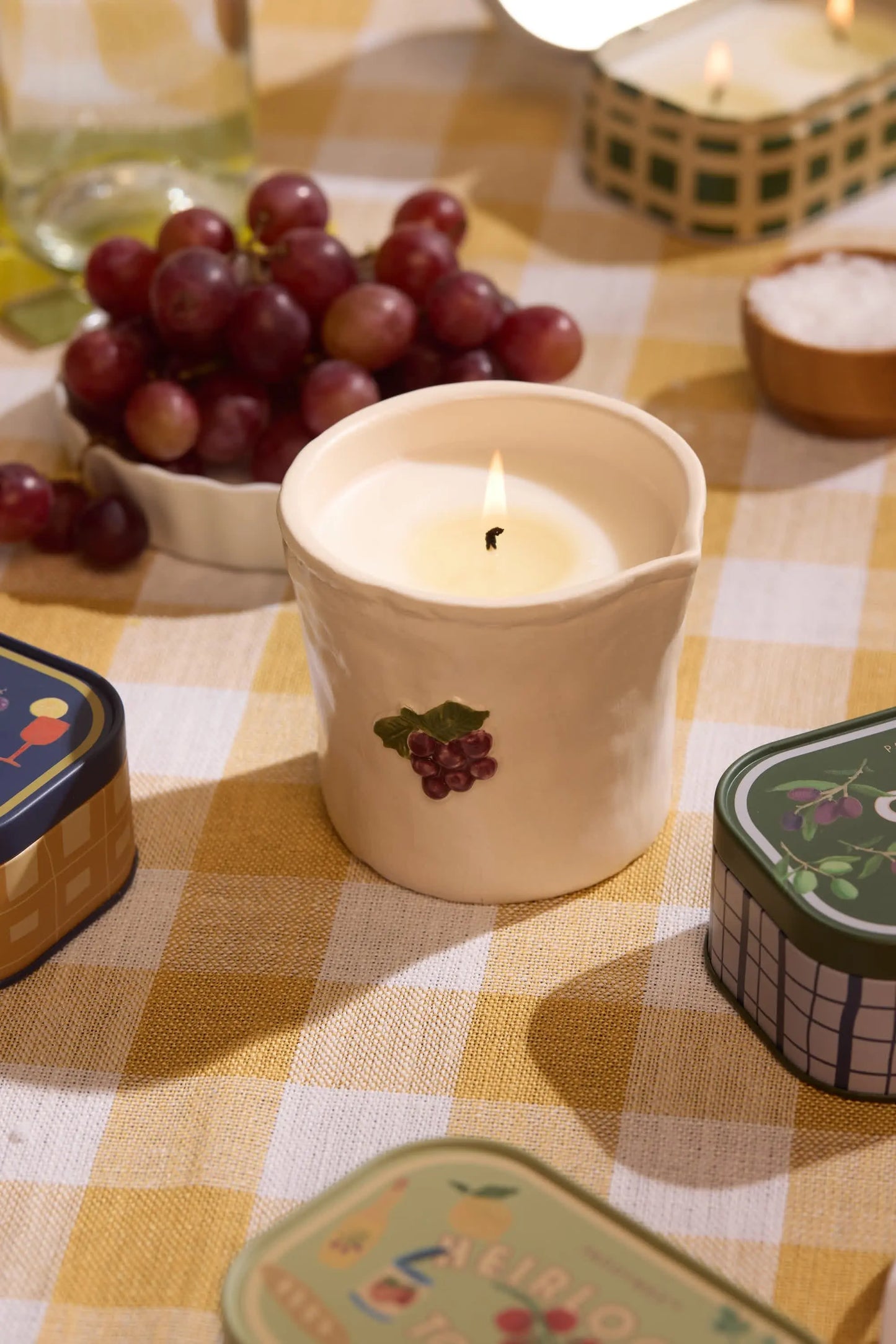 A Bistro Candle - Bordeaux Grape in a white ceramic holder with grape design sits on a yellow checkered tablecloth, surrounded by grapes and tins, capturing the charm of culinary culture.