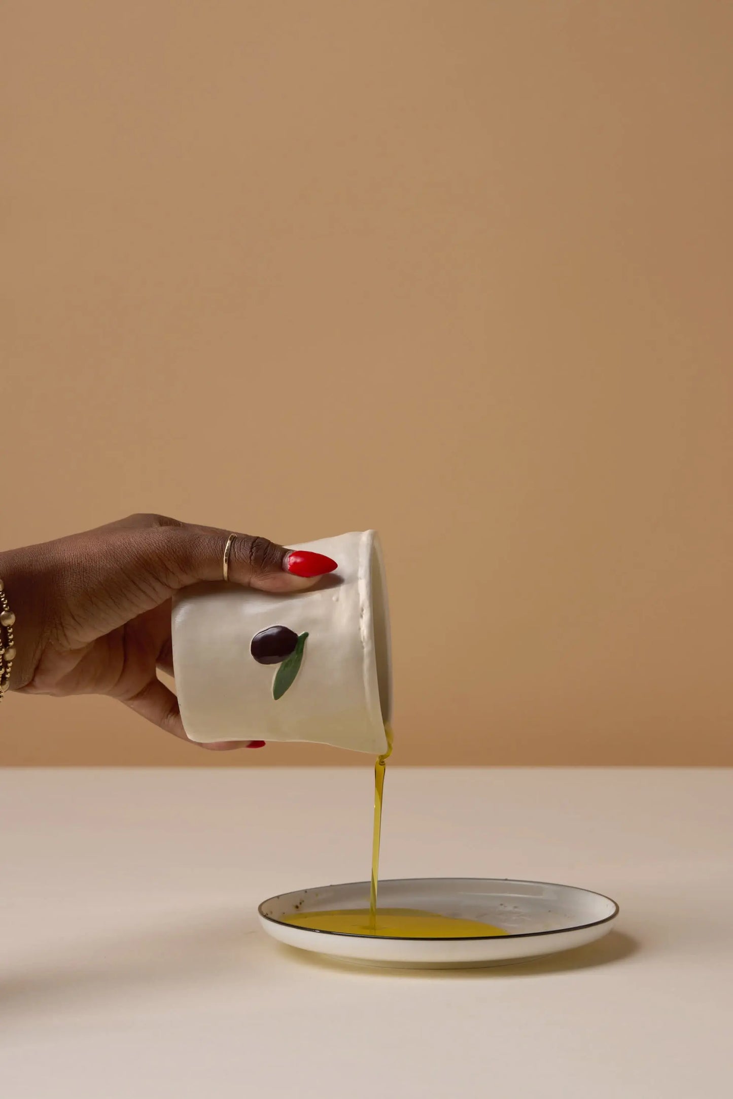 A hand with red-painted nails pours olive oil from a white cup with hand-painted food icons onto a dish, featuring the Fig & Olive - Bistro Candle 8 oz, against a beige background.