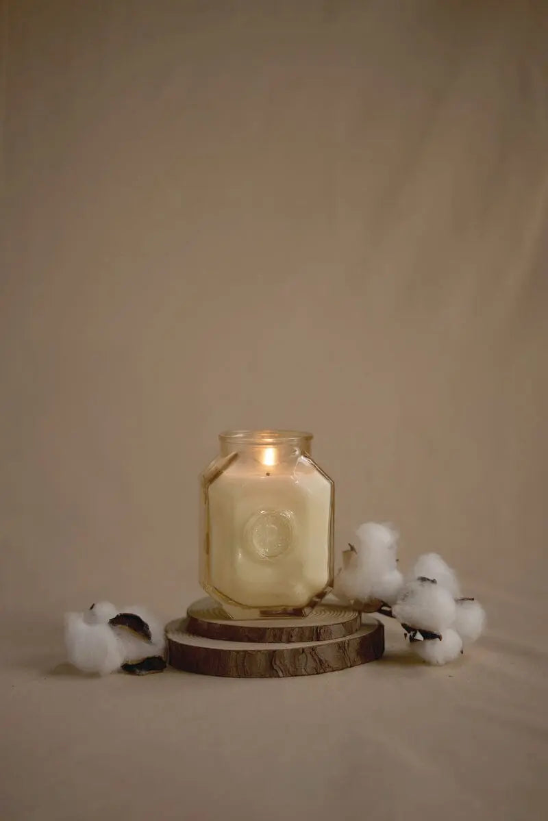 The Botanica Candle - Cotton + Teak from the Luxurious Botanica Collection glows in a glass jar atop stacked wooden slices, with cotton stems nearby, all set against a plain beige background.