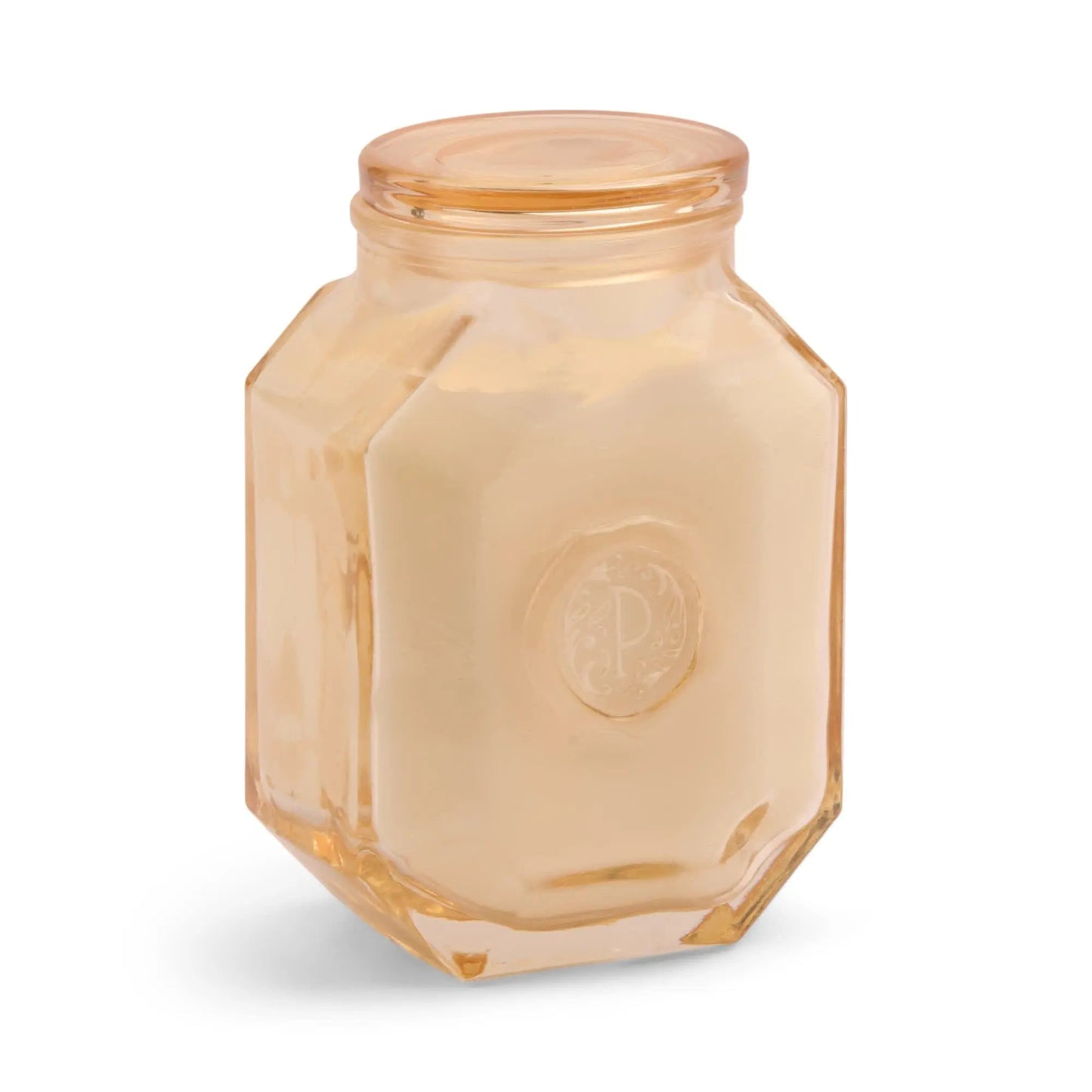 The Botanica Candle - Cotton + Teak features a hexagonal glass jar with a light peach tint and cream-colored wax, sealed with a matching lid—ideal for adding elegance to your décor.