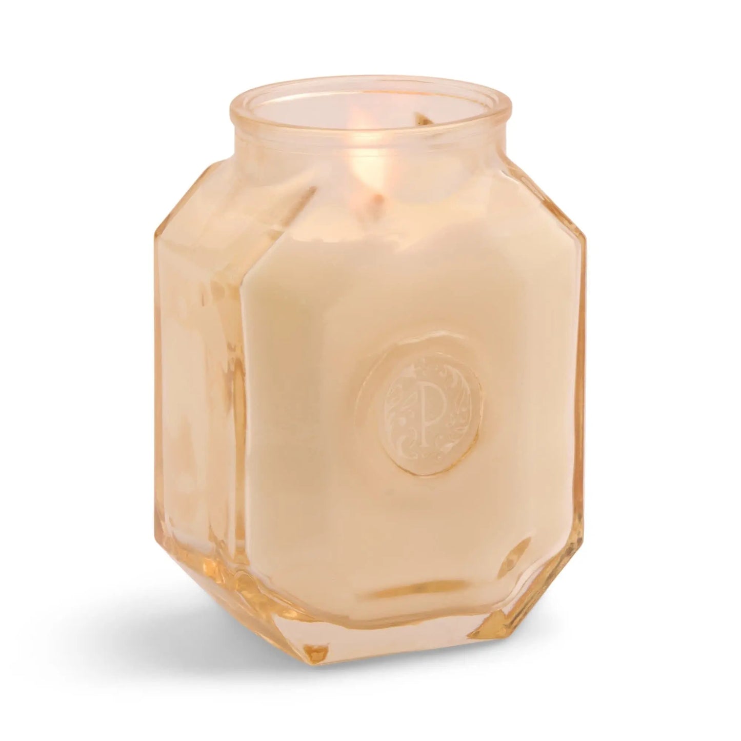 The Botanica Candle - Cotton + Teak glows in a translucent peach hexagonal jar adorned with a decorative emblem—an elegant accent to your candles and diffusers display.