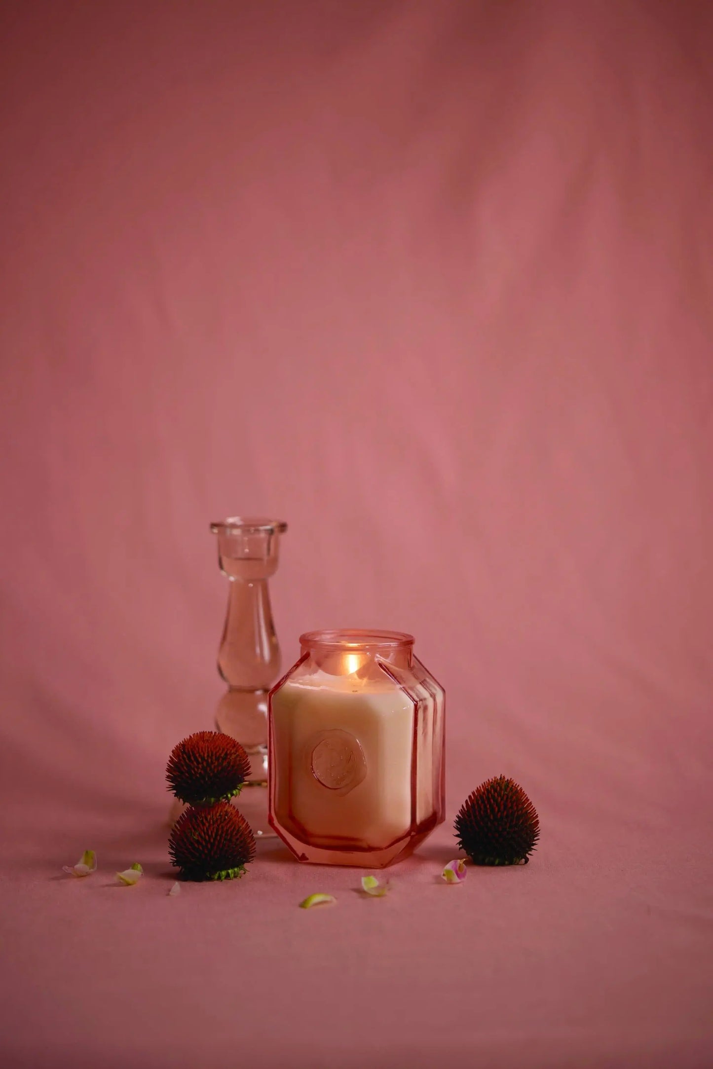 A lit Botanica Candle - Saffron Rose in a pink glass jar sits beside a clear candle holder and three round textured objects on a pink surface with a pink background.