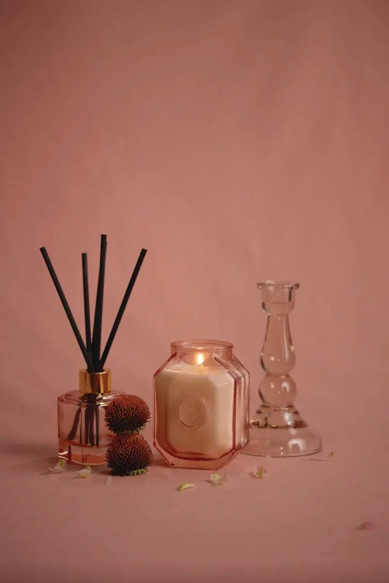 The Botanica Glass Diffuser - Saffron Rose with black reeds, a lit candle in a pink jar, two dried flowers, and an empty glass candlestick holder are elegantly arranged on a pink surface.